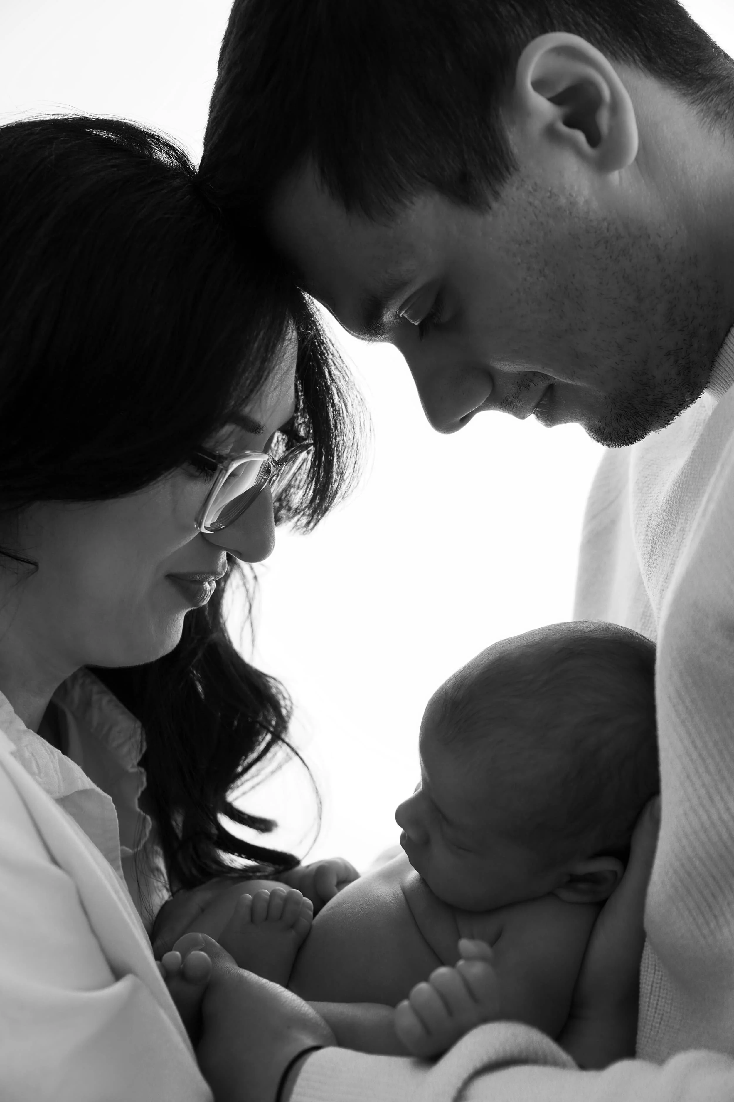 Ein Schwarz-Weiß-Foto einer Familie, die ein Neugeborenes liebevoll hält, mit den Köpfen nah beieinander und Blicken auf das Baby gerichtet.Emotionale Studiofotografie Babybauch und Neugeborene in Schwenningen und Umgebung.