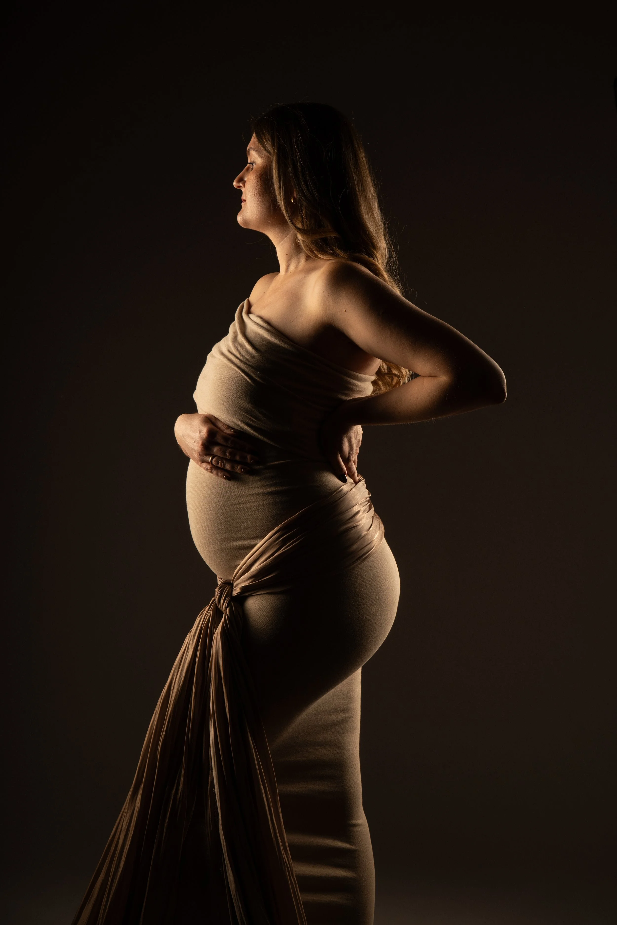 Seitenansicht einer schwangeren Frau, in einem engen, beigen Stoff, der die Schwangerschaft edel betont, vor dunklem Hintergrund. KaysserArt Fotografie
