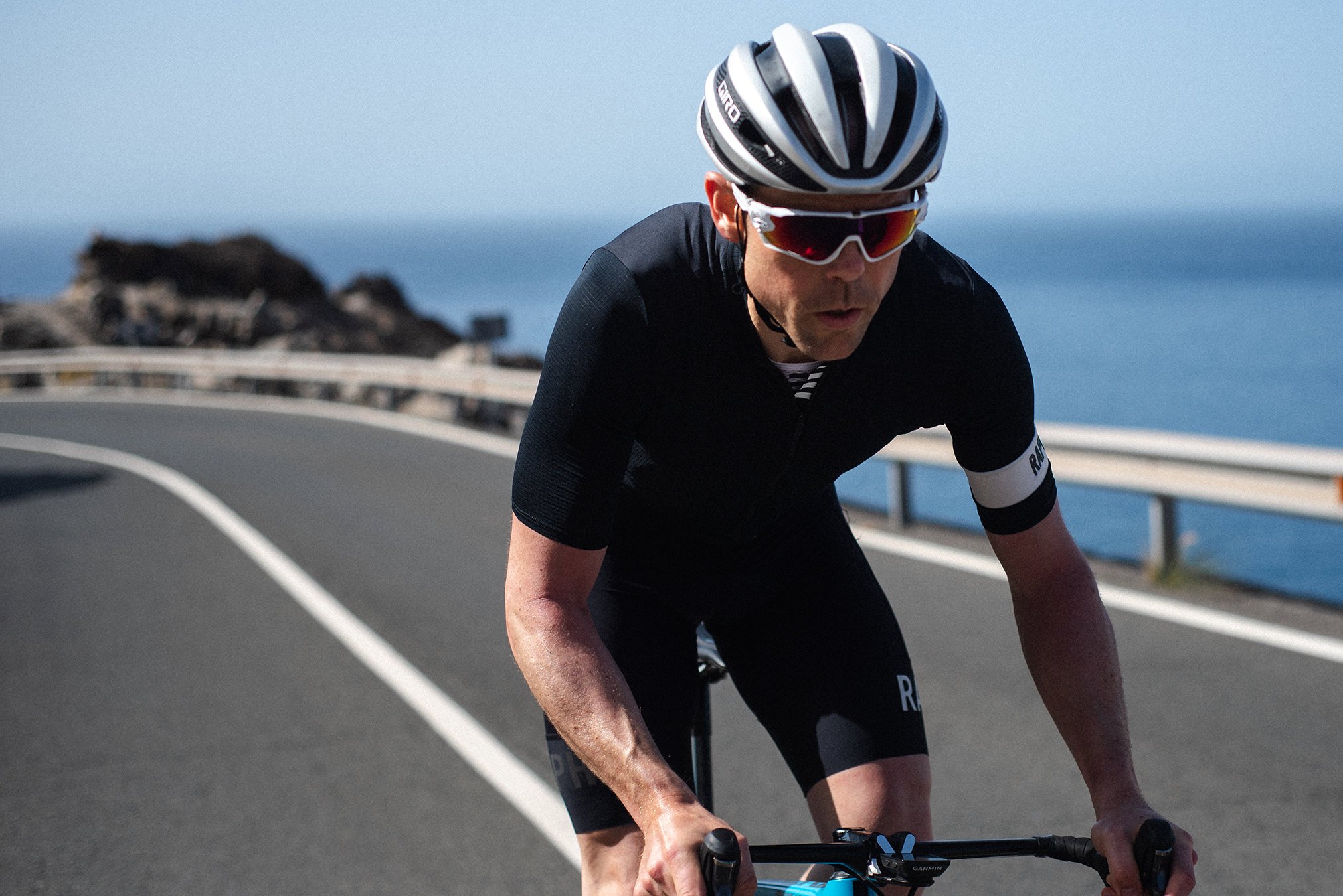 A male cyclist wearing a helmet and sunglasses rides a bicycle on a coastal road.