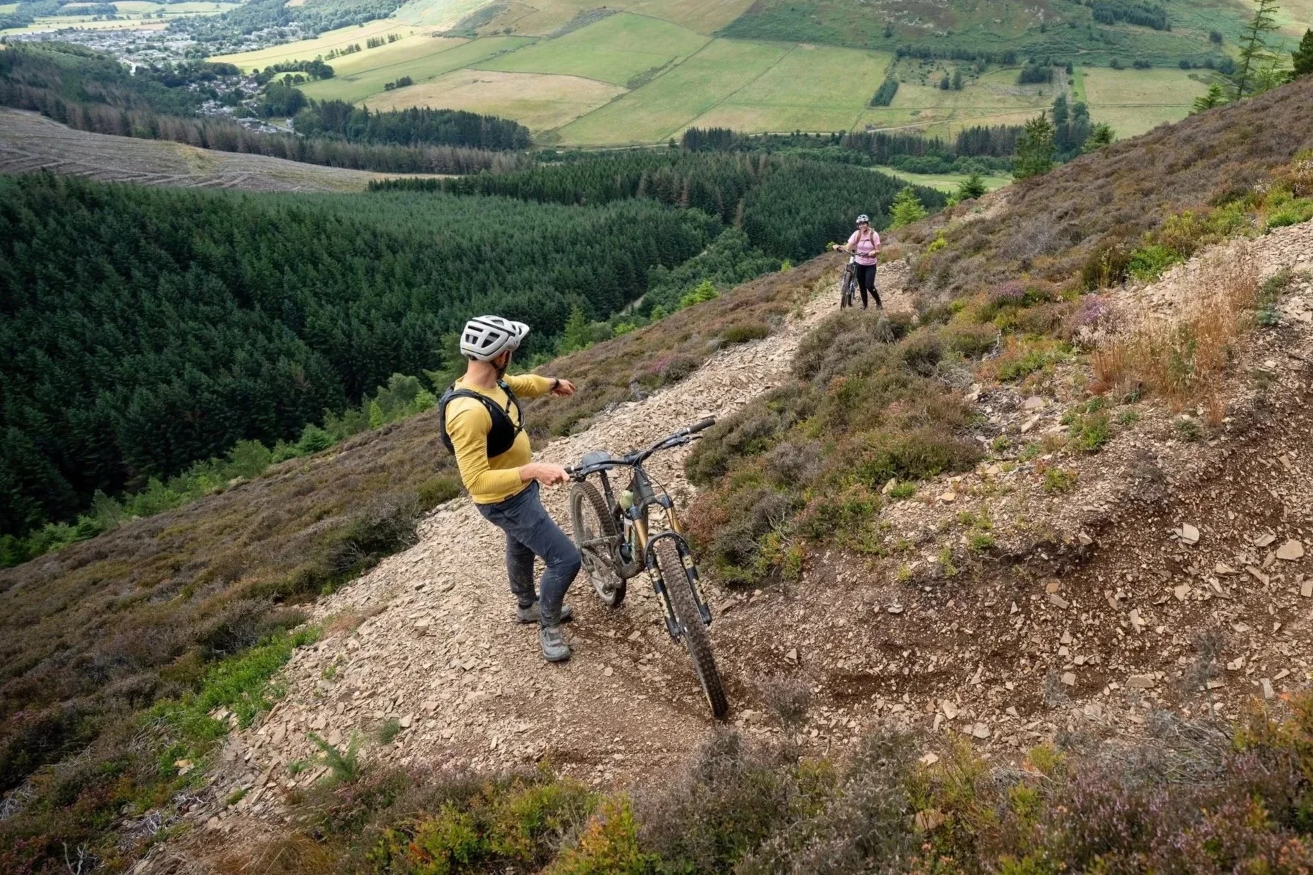 A person in a yellow long-sleeve shirt and gray pants holding a mountain bike on a rocky trail on a hillside, with another person in a pink shirt and black pants standing further along the trail with a mountain bike, overlooking a green valley with patchwork fields, forests, and distant hills.