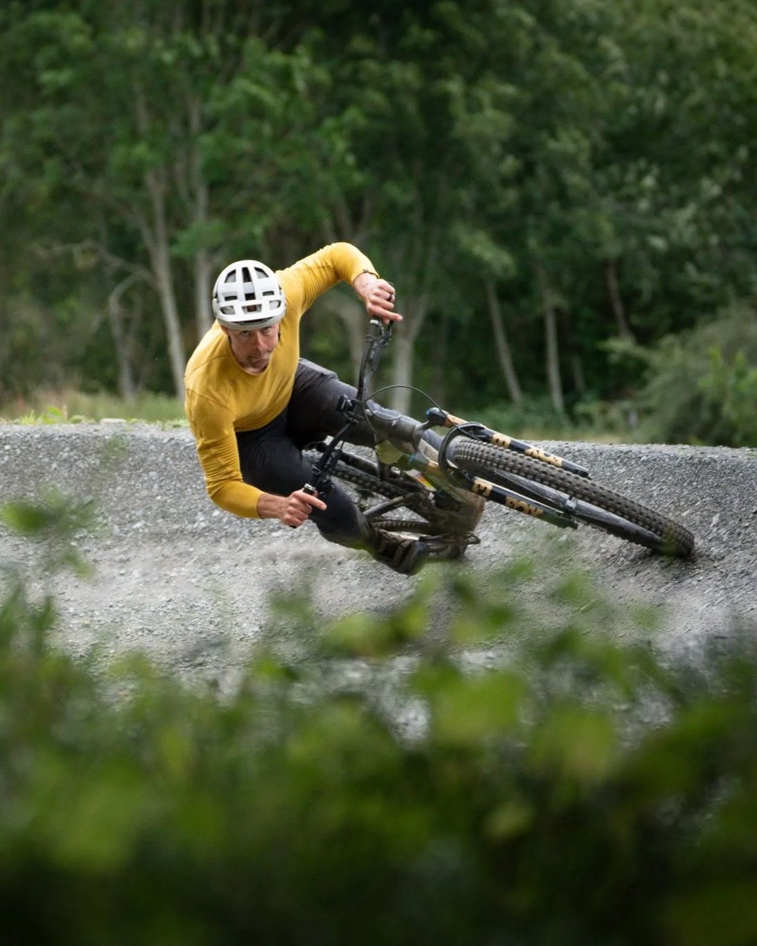 A person wearing a white helmet and a yellow long-sleeve shirt is mountain biking on a gravel trail through a forest, leaning sharply into a turn.