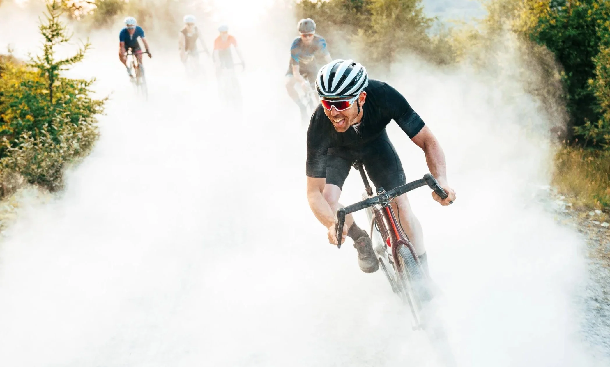 A group of mountain bikers riding through a foggy or dusty trail in a forested area, with one biker in the foreground wearing a helmet and sunglasses, smiling while riding.