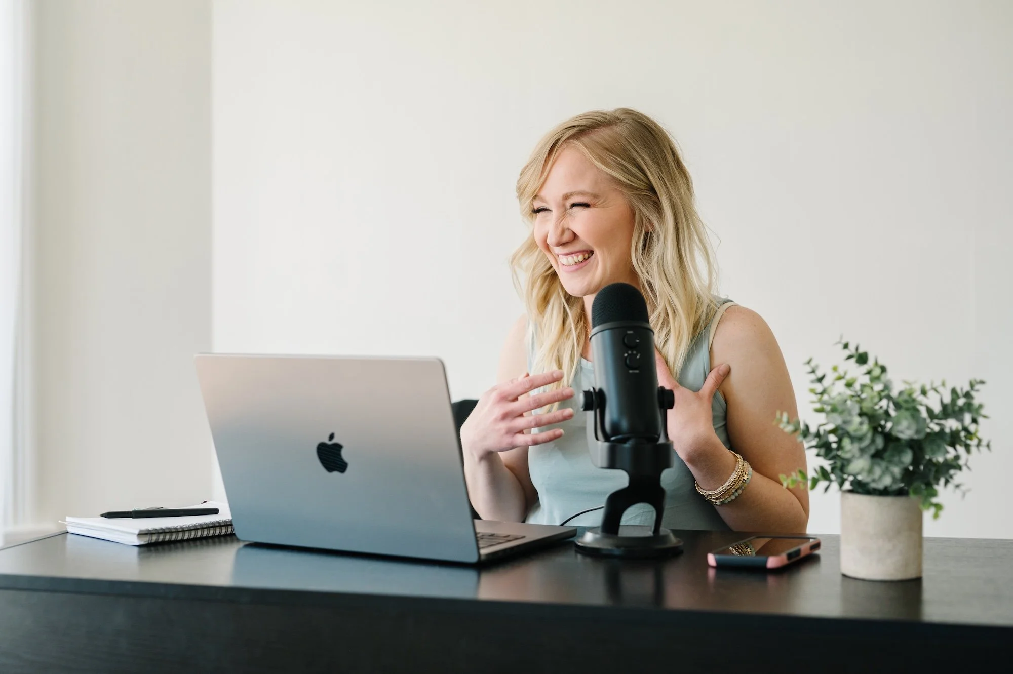 A woman, Megan Lloyd, with blonde hair smiling and laughing in front of a laptop with a microphone on a desk.  She has helped 175+ clients around the globe hire virtual assistants and social media managers. 