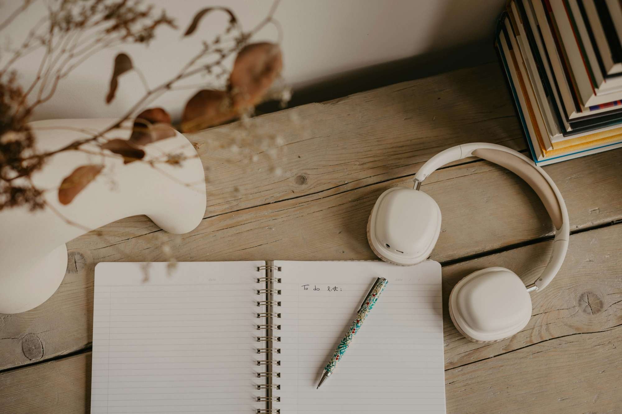 A wooden desk with a white spiral notebook open to a blank page labeled 'To do list:', a floral pen, white over-ear headphones, a white ceramic vase with dried flowers, and a stack of files or magazines.