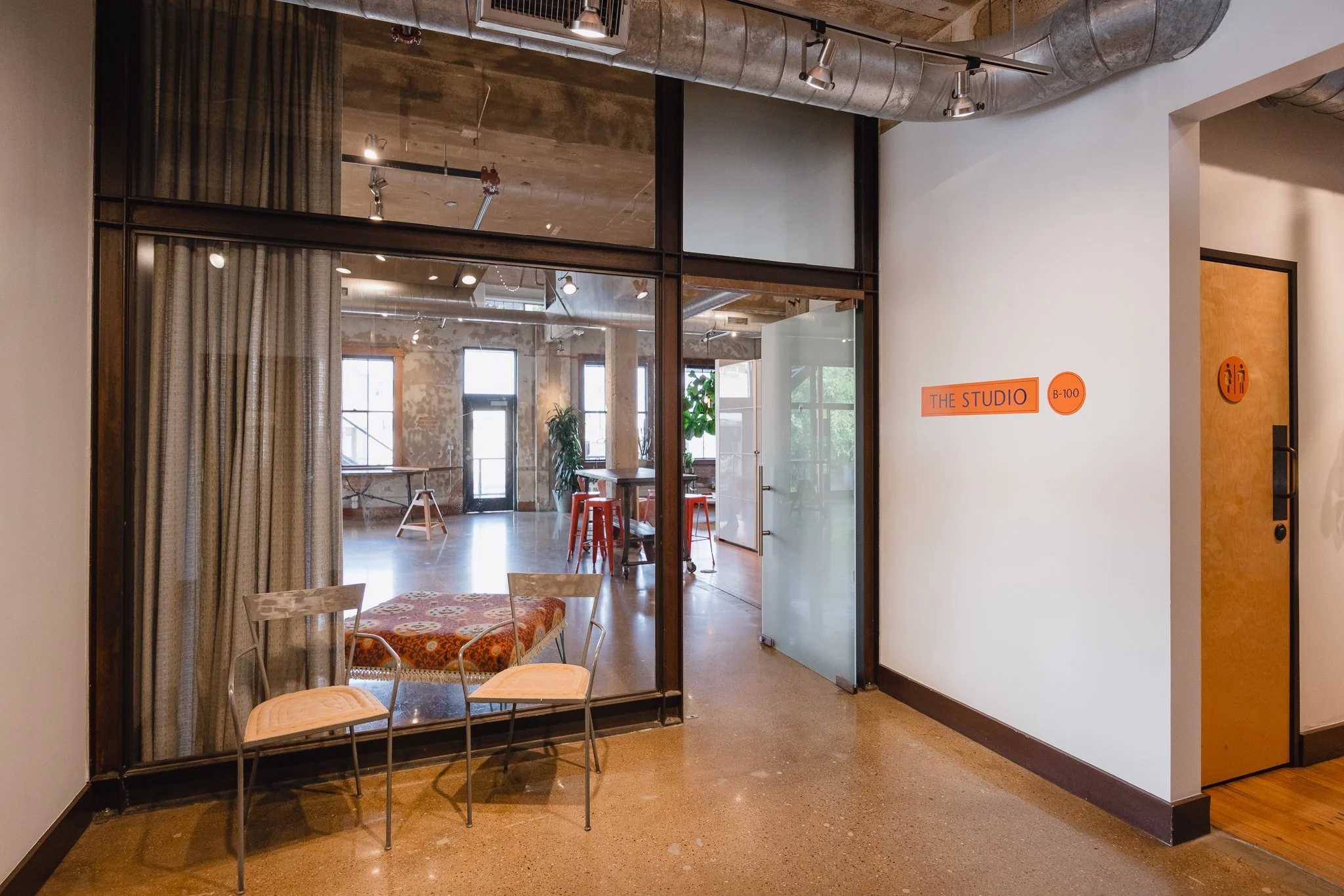 Interior view of a modern studio space seen through a large glass window and door, with a small sitting area in the foreground featuring two chairs and a table, and an open room with high windows, exposed brick walls, plants, and furniture in the bac