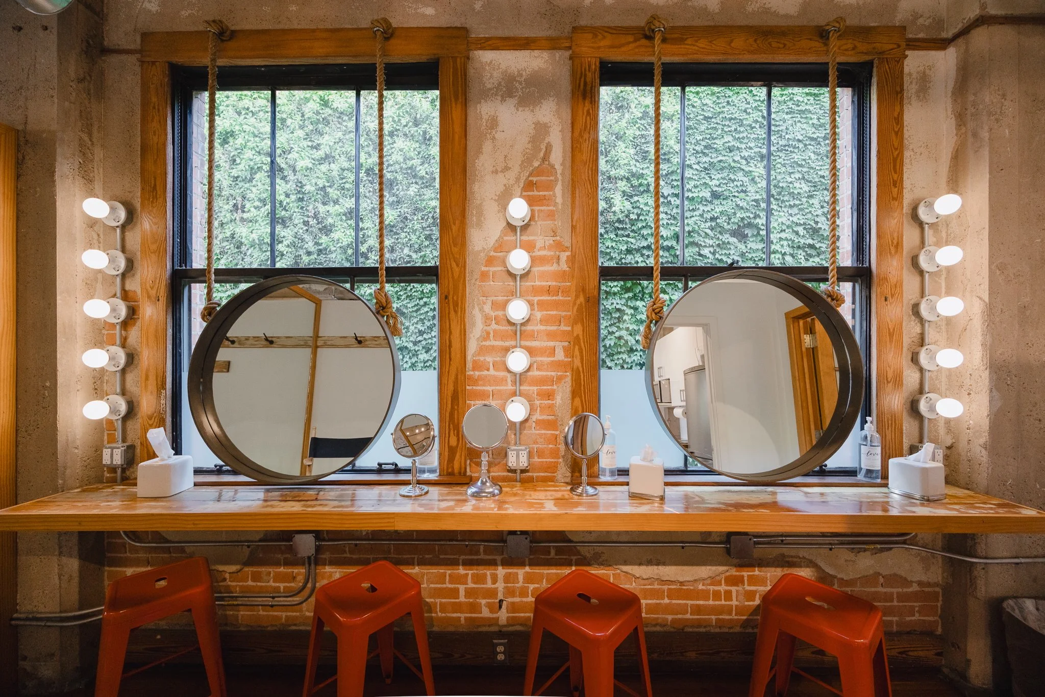 A stylish salon station with two round mirrors hanging from ropes in front of large windows with view of greenery. The wooden countertop holds small mirrors, tissue boxes, and hand sanitizer. Four red stools are placed in front of the counter.