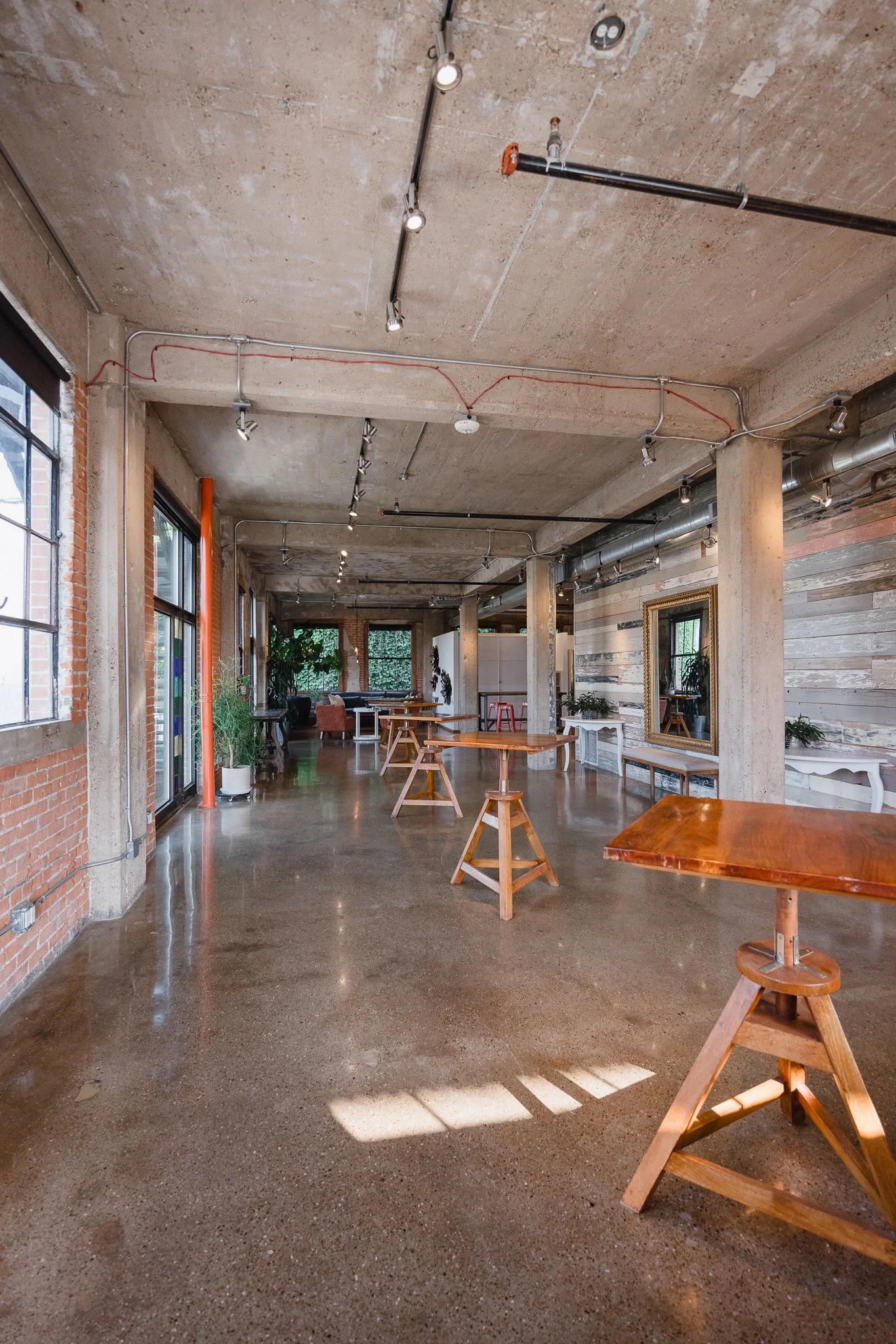 An industrial-style interior space with wooden tables, concrete ceiling, exposed pipes, large windows, potted plants, and wall decor.