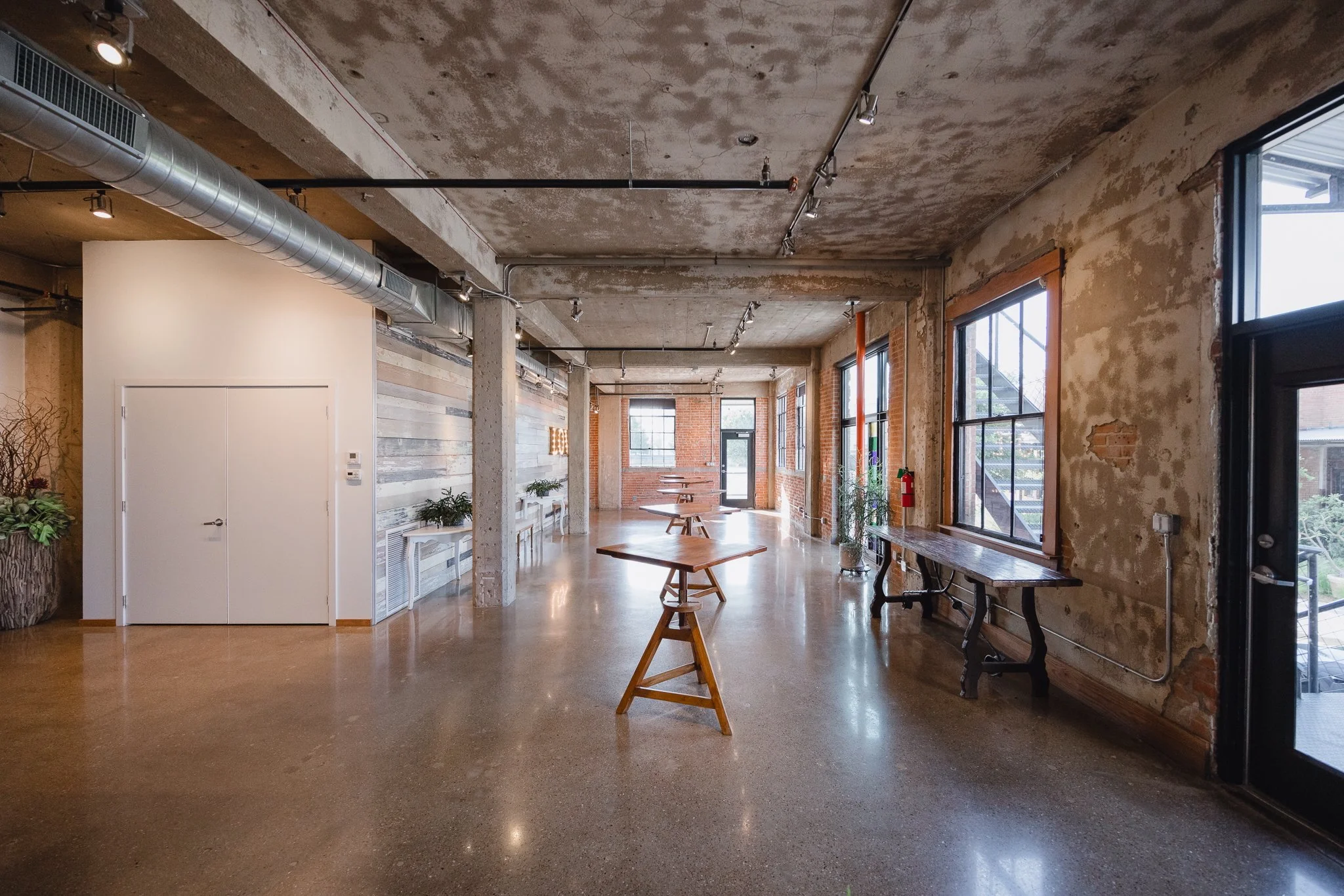 Interior of an industrial-style open space with exposed concrete ceiling, large windows, wooden accents, and a polished concrete floor. Contains wooden tables and potted plants.