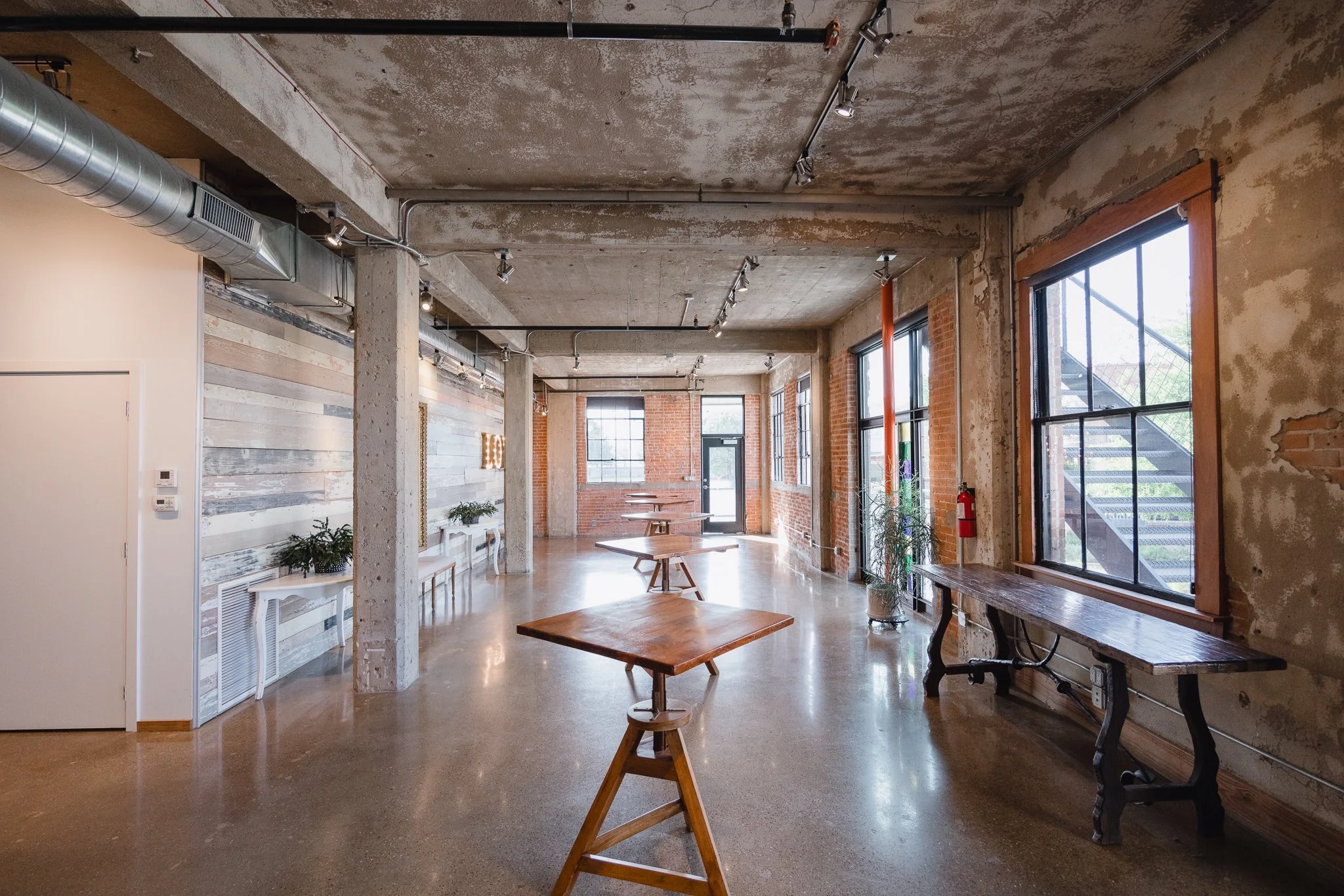 An industrial-style interior space with exposed concrete ceilings, brick walls, large windows, and wooden tables.