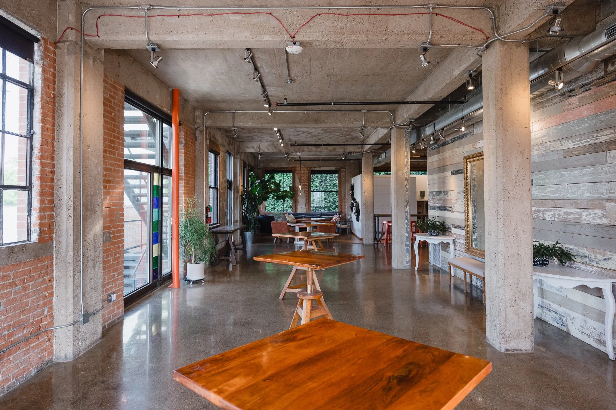 Interior of a modern, industrial-style cafe with wooden tables, exposed brick and concrete walls, large windows, green plants, and track lighting.