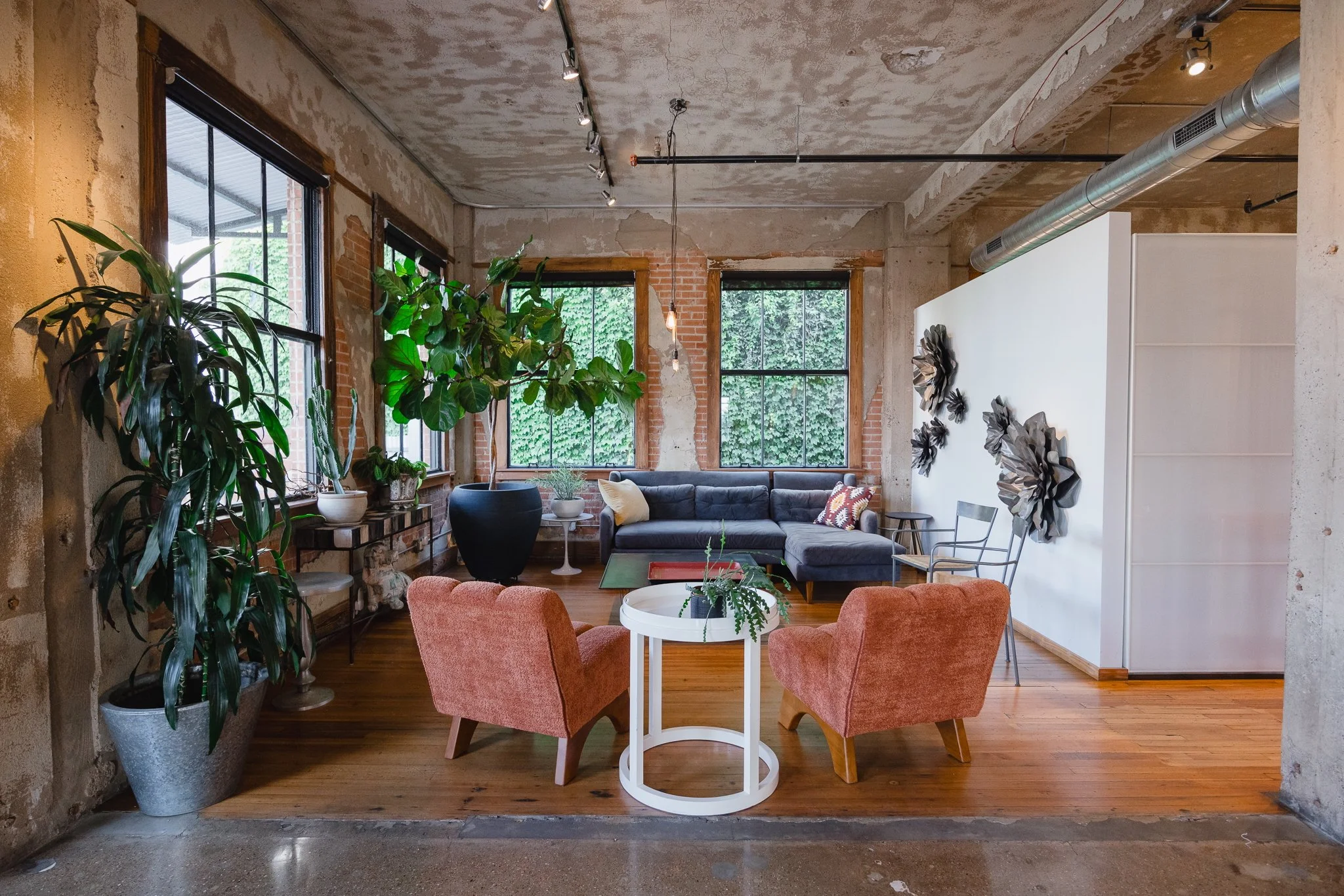 A cozy, industrial-style living room with exposed brick and concrete walls, large windows with black frames, and a mix of modern and vintage furniture. It features a dark sectional sofa, two rust-colored armchairs around a white round side table with