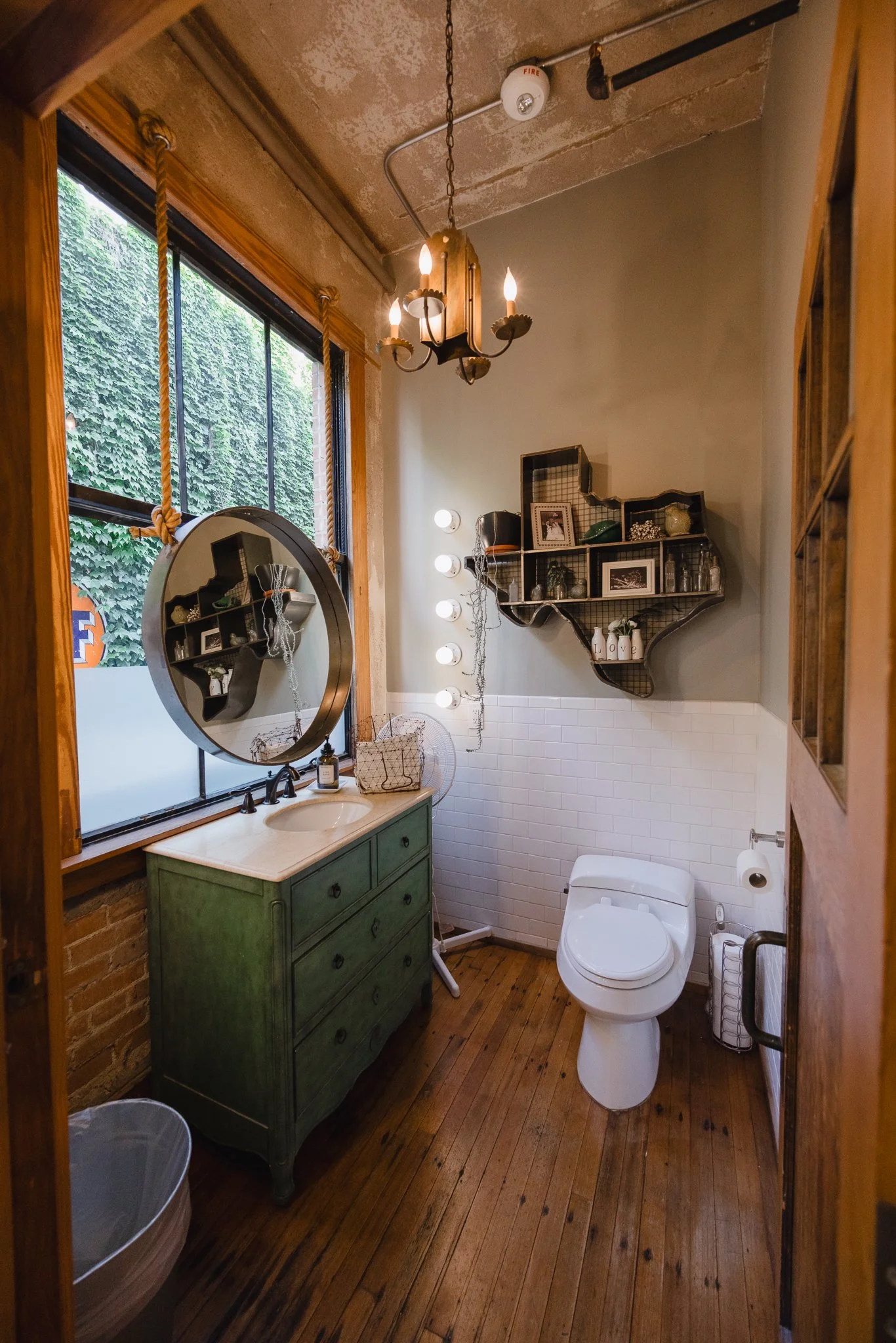 A small bathroom with wooden floors, a green dresser with a marble top, a round mirror, a window with wooden trim, a modern toilet, a wire wall shelf with decorative items, and a chandelier with candles hanging from the ceiling.