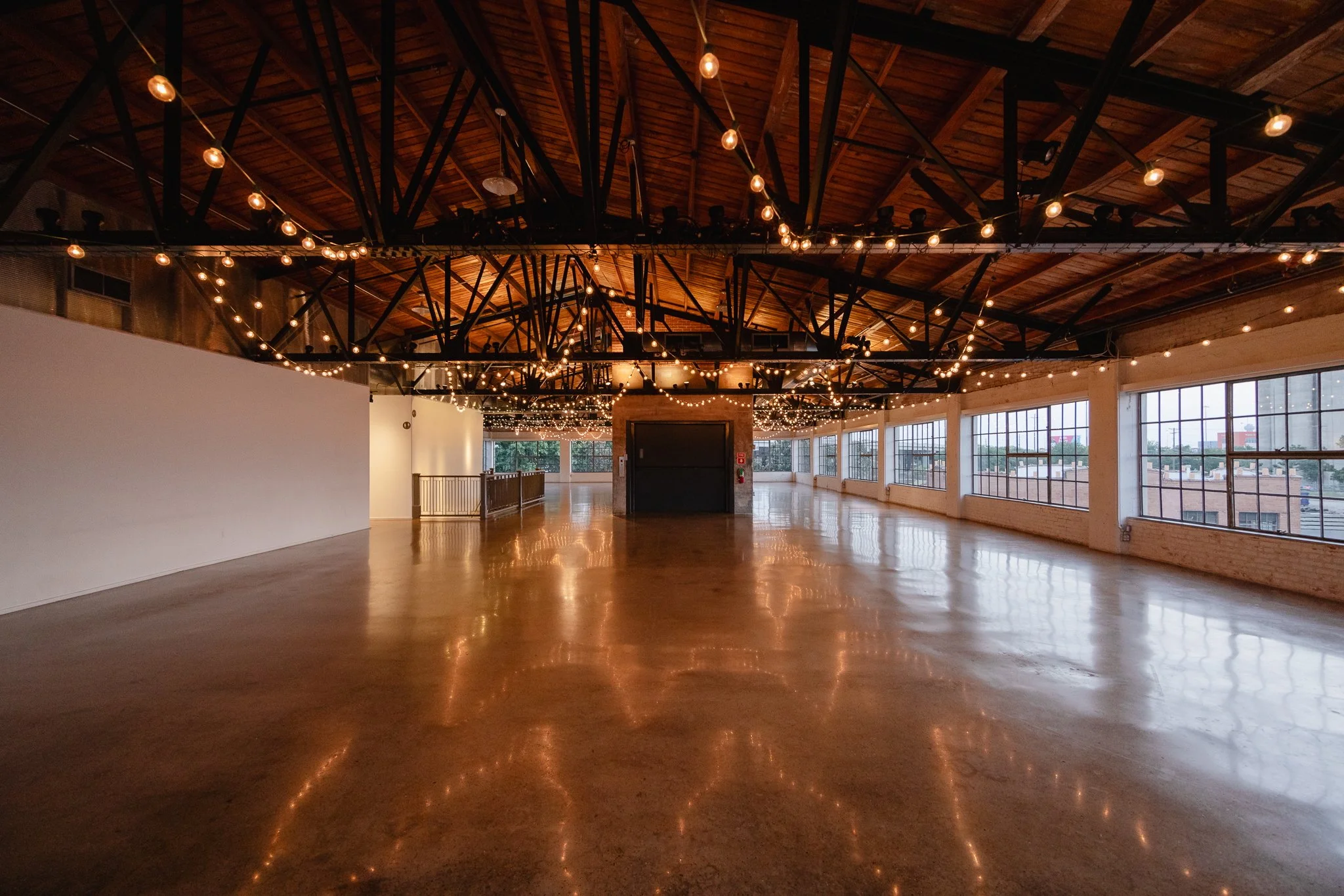 Empty event space with wooden ceiling, string lights, large windows, and polished concrete floor, decorated for a gathering or celebration.