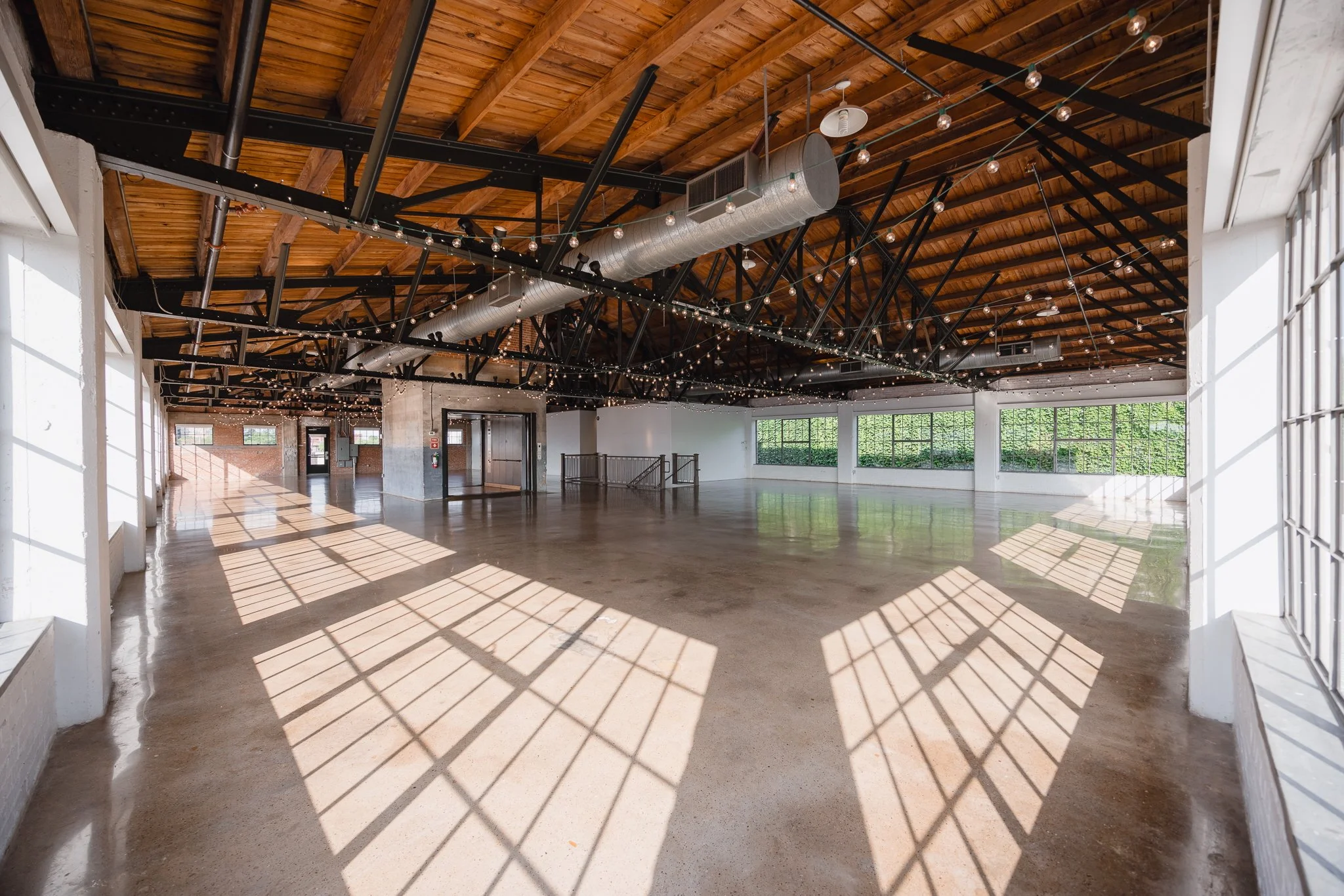 Empty industrial-style event space with large windows, polished concrete floor, wooden ceiling with exposed beams, string lights, and outdoor greenery visible through windows.