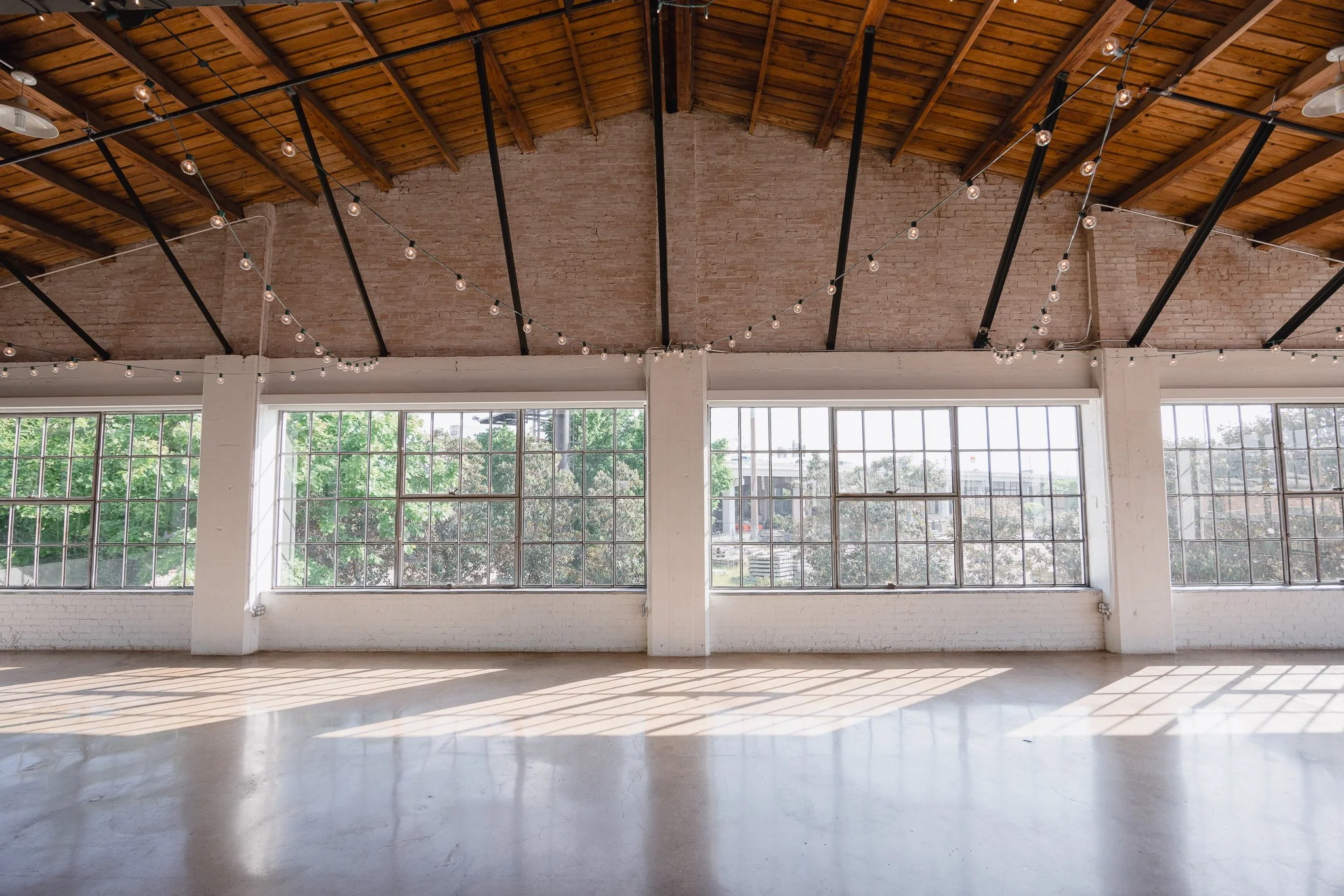 An empty industrial-style room with large windows, wooden ceiling, and string lights hanging from the ceiling.