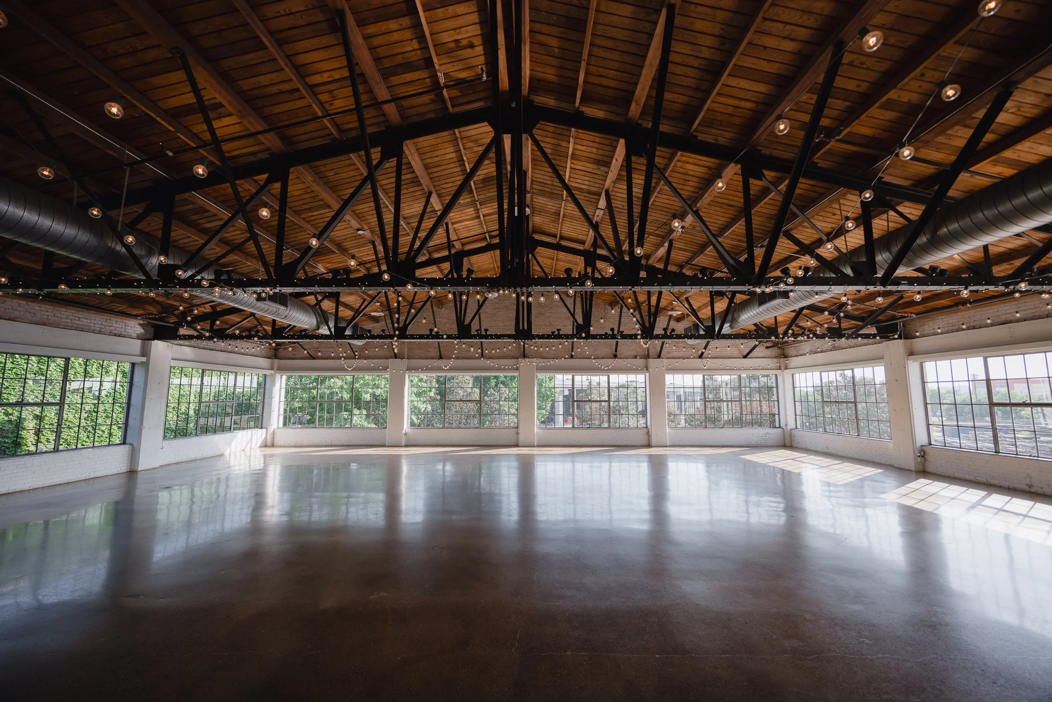 Empty event space with large windows, polished concrete floors, exposed brick walls, and string lights hanging from the high wooden ceiling.