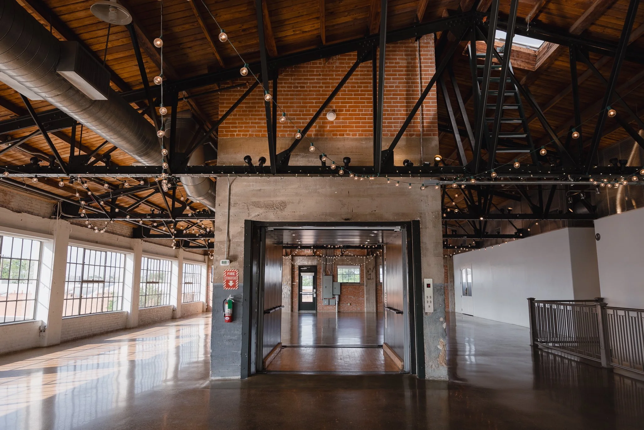 An industrial-style interior space with wooden ceiling, black metal beams, string lights, large windows, a brick and concrete wall, an elevator with open doors, and a polished concrete floor.