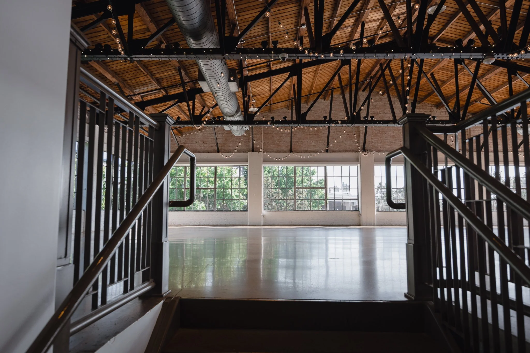 Empty indoor event space with large windows, exposed brick walls, a polished floor, string lights hanging from the wooden ceiling, and black metal railings.