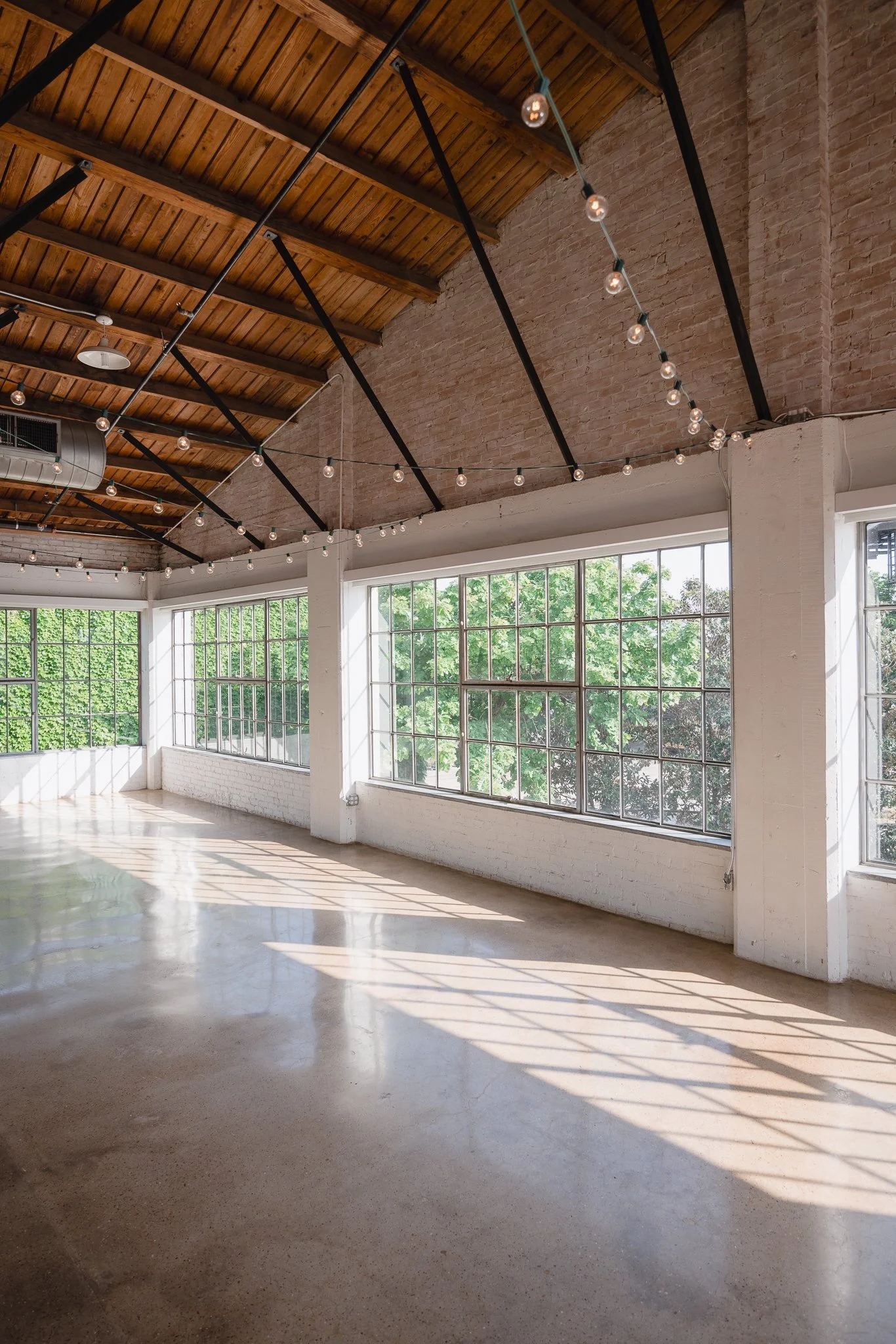 Bright, spacious indoor area with large windows, exposed brick walls, polished concrete floor, and string lights hanging from a wooden ceiling.
