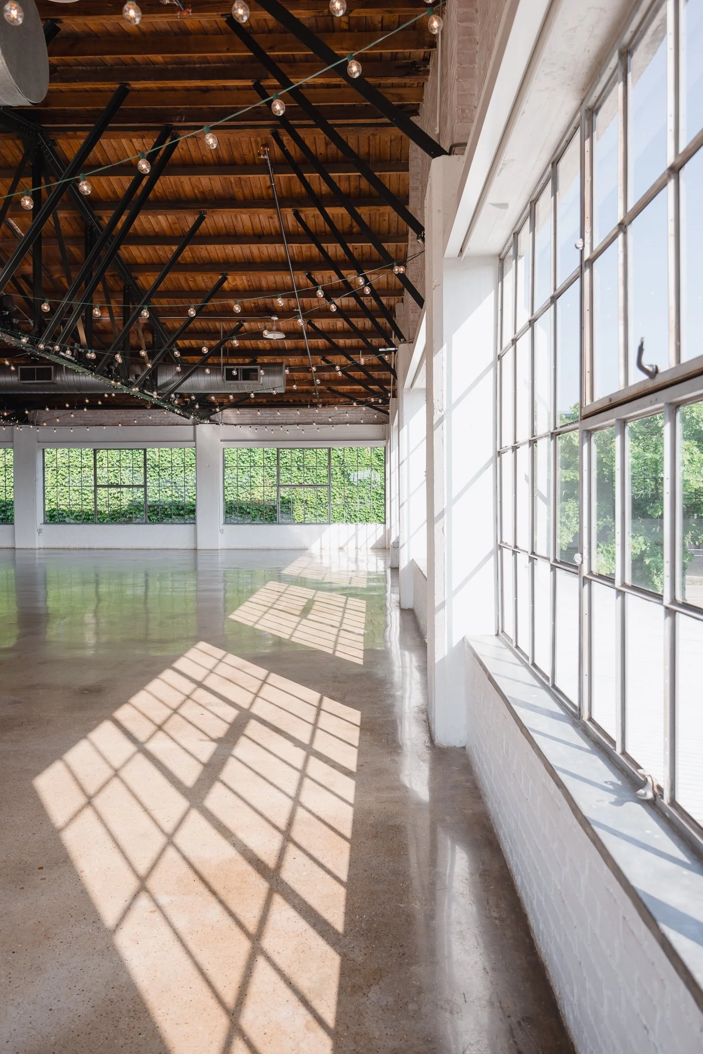 Bright indoor space with large windows letting in sunlight, wooden ceiling beams, string lights, polished concrete floor, and greenery outside.