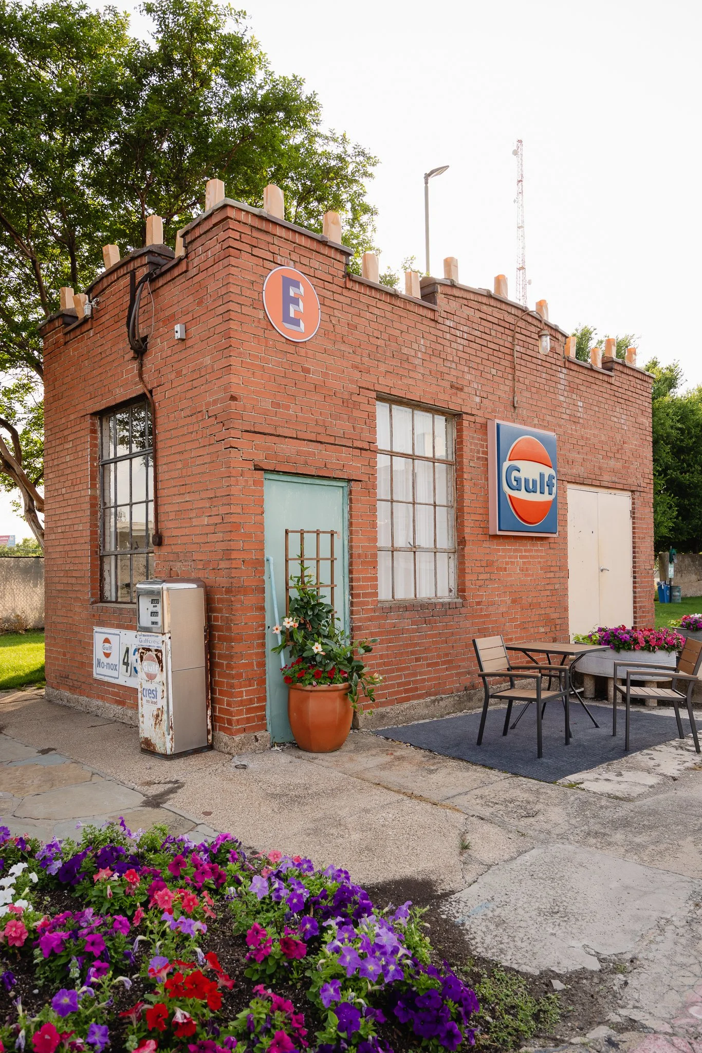 A small brick building with Gulf signage, a blue door with a potted plant, outdoor seating, and colorful flowers in the foreground.