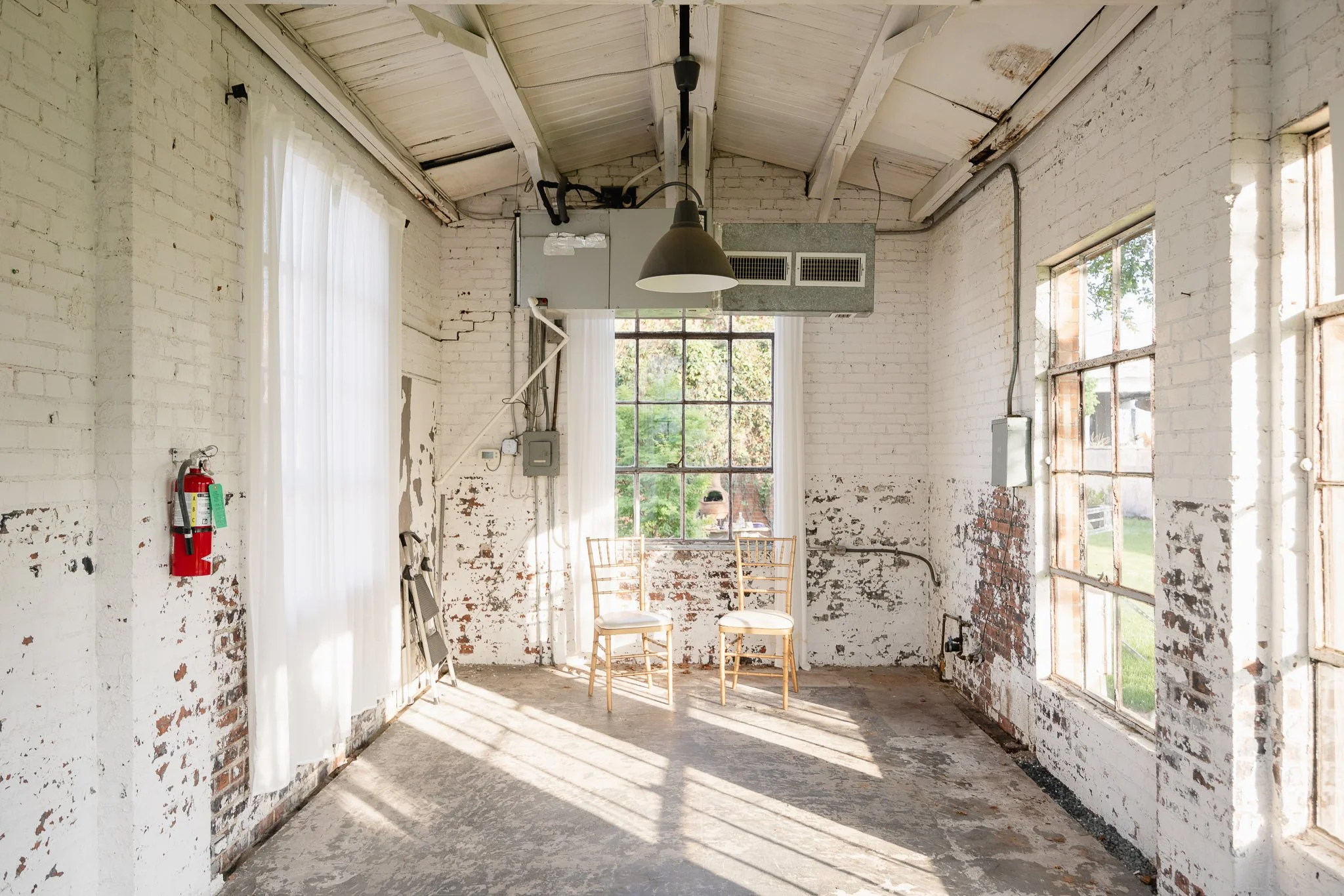 Empty room with white brick walls, a large window with sheer curtains, two chairs, a fire extinguisher on the wall, and an industrial-style hanging light fixture.
