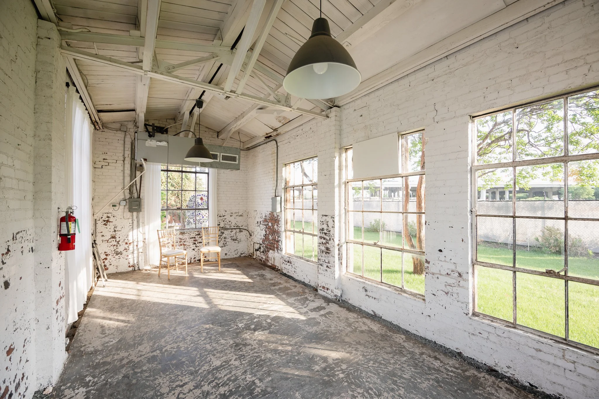 Empty industrial-style room with white brick walls, large windows, hanging pendant lights, and two chairs near the window.