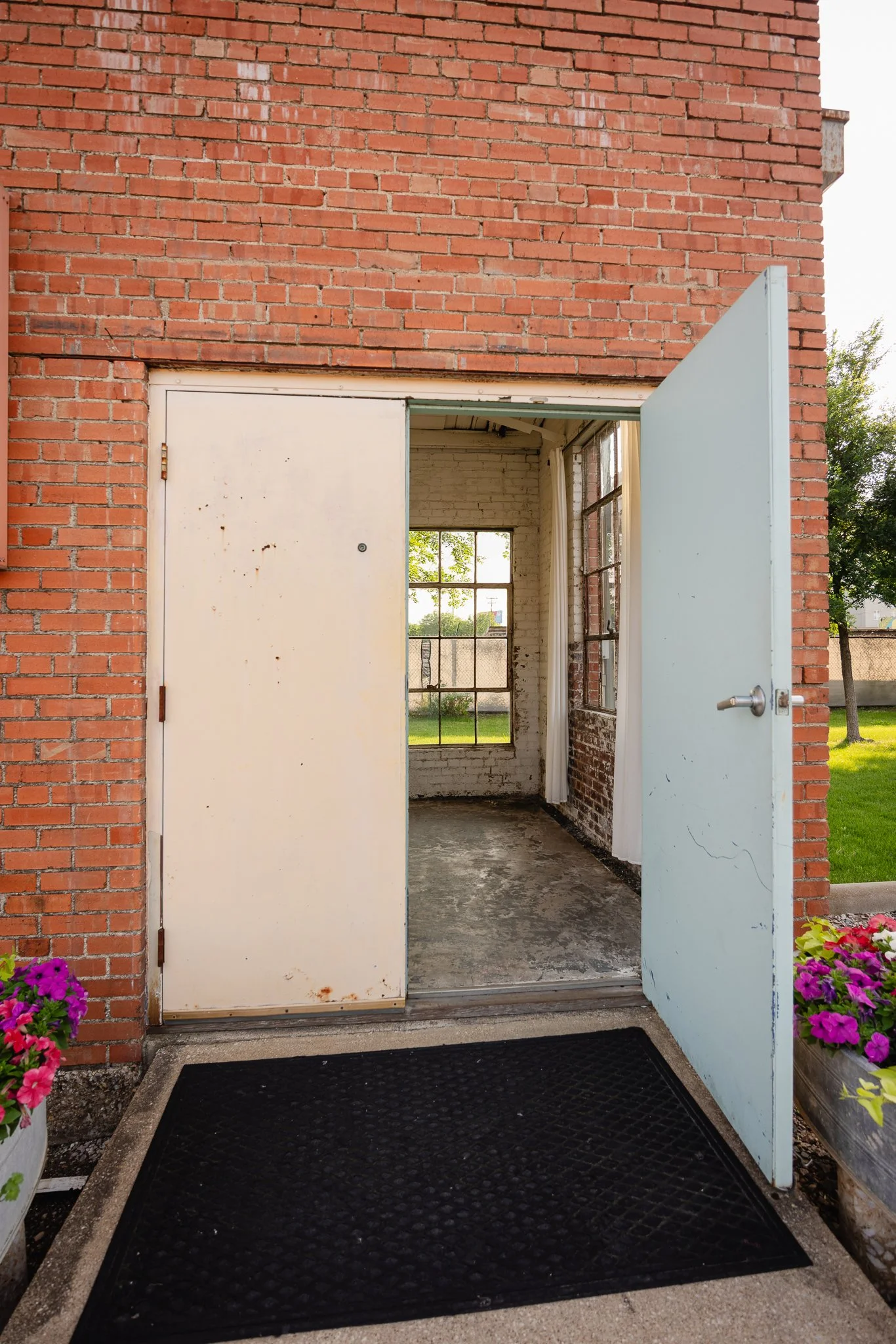 Open door leading into a rustic interior with brick walls and large window, outside green grass and trees, flower planters on either side of the entrance.
