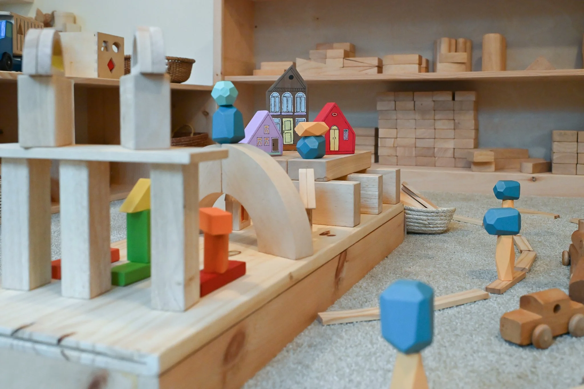 A thoughtfully designed block area has a large collection of wooden blocks of various shapes;  it is accessorized with colorful toy houses, trees, cars, trains and a collection of toy animals.