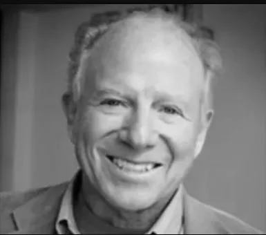 Black and white photo of a smiling middle-aged man with short, light-colored hair, wearing a suit jacket and collared shirt.
