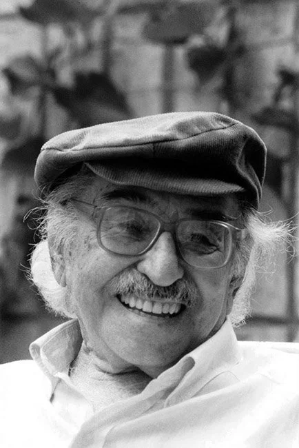 Older man with glasses, a cap, and a mustache smiling in front of a blurred natural background.