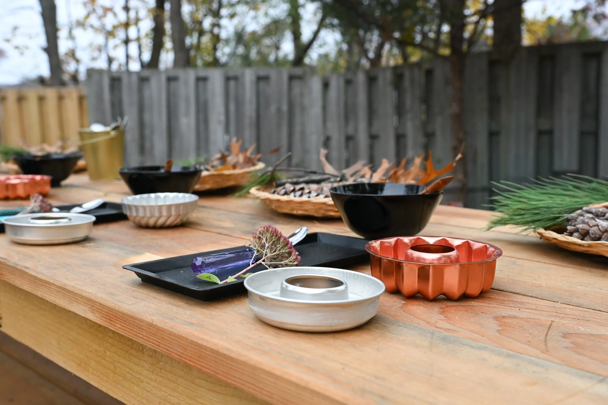 An outdoor table offers rich opportunities for play and explorations with various containers, dishes and kitchen utensils.  The mud kitchen is filled with plenty of natural elements like mud, clay, foliage, pinecones, tree cookies and sticks.