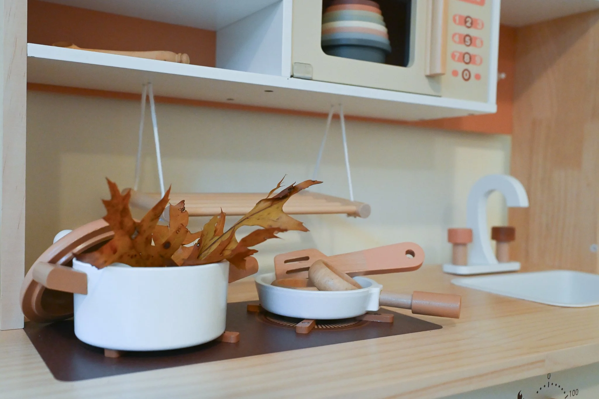 A toy kitchen set including a white pot with leaves, a small white pan with a wooden spoon, and a white faucet, with toy dishes and utensils on a wooden countertop.