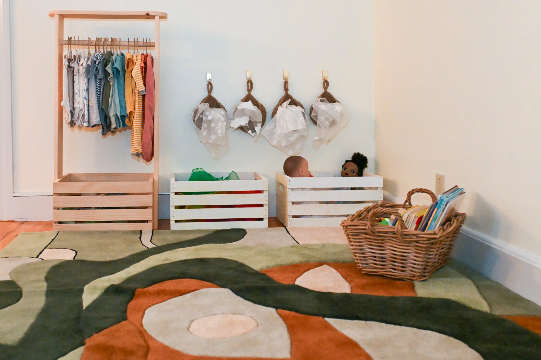 Children's playroom with a wooden clothing rack holding folded and hanging clothes, four paper bags hanging on hooks, two dolls in a white wooden crate, a woven basket with books, and a colorful area rug with abstract patterns.