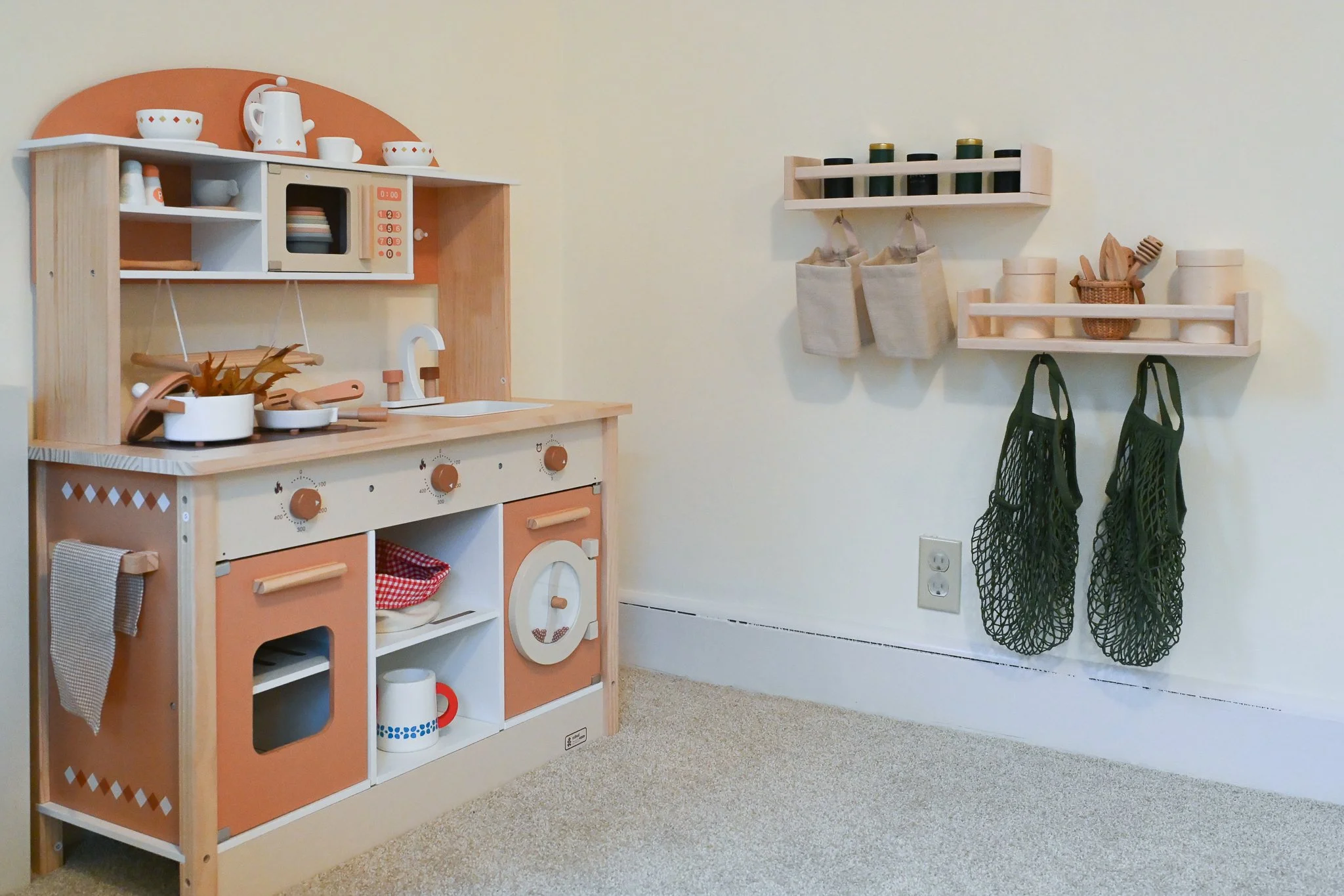 Children's wooden play kitchen with accessories, wall shelves with containers and bags, and green mesh bags hanging, beige carpeted floor.