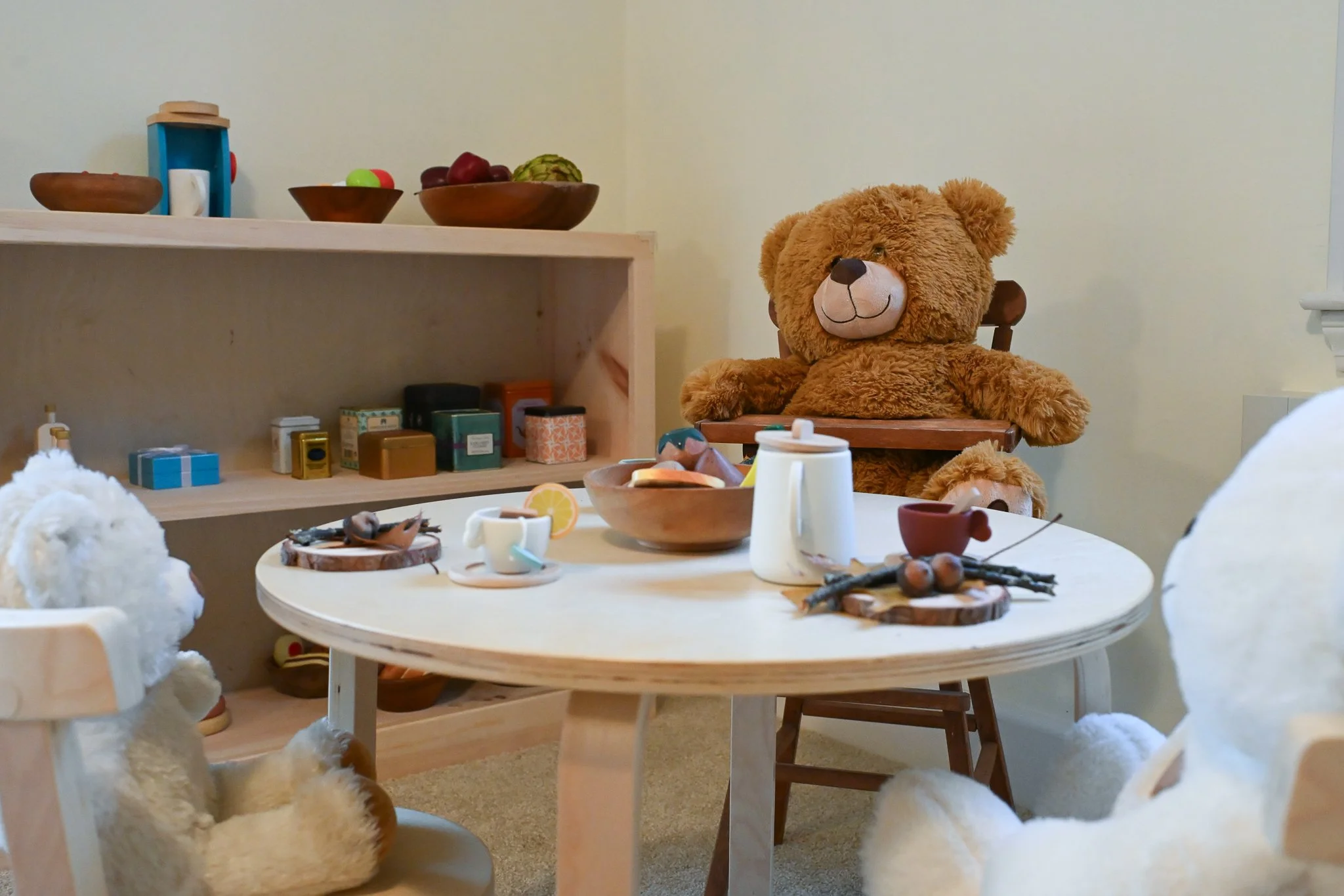 Children's dramatic play area with a thoughtful collection of toys and materials for pretend play. Round wooden table set with toy teacups, a bowl with toy food, and a white pitcher. Plush teddy bears and stuffed animals are seated around the table.