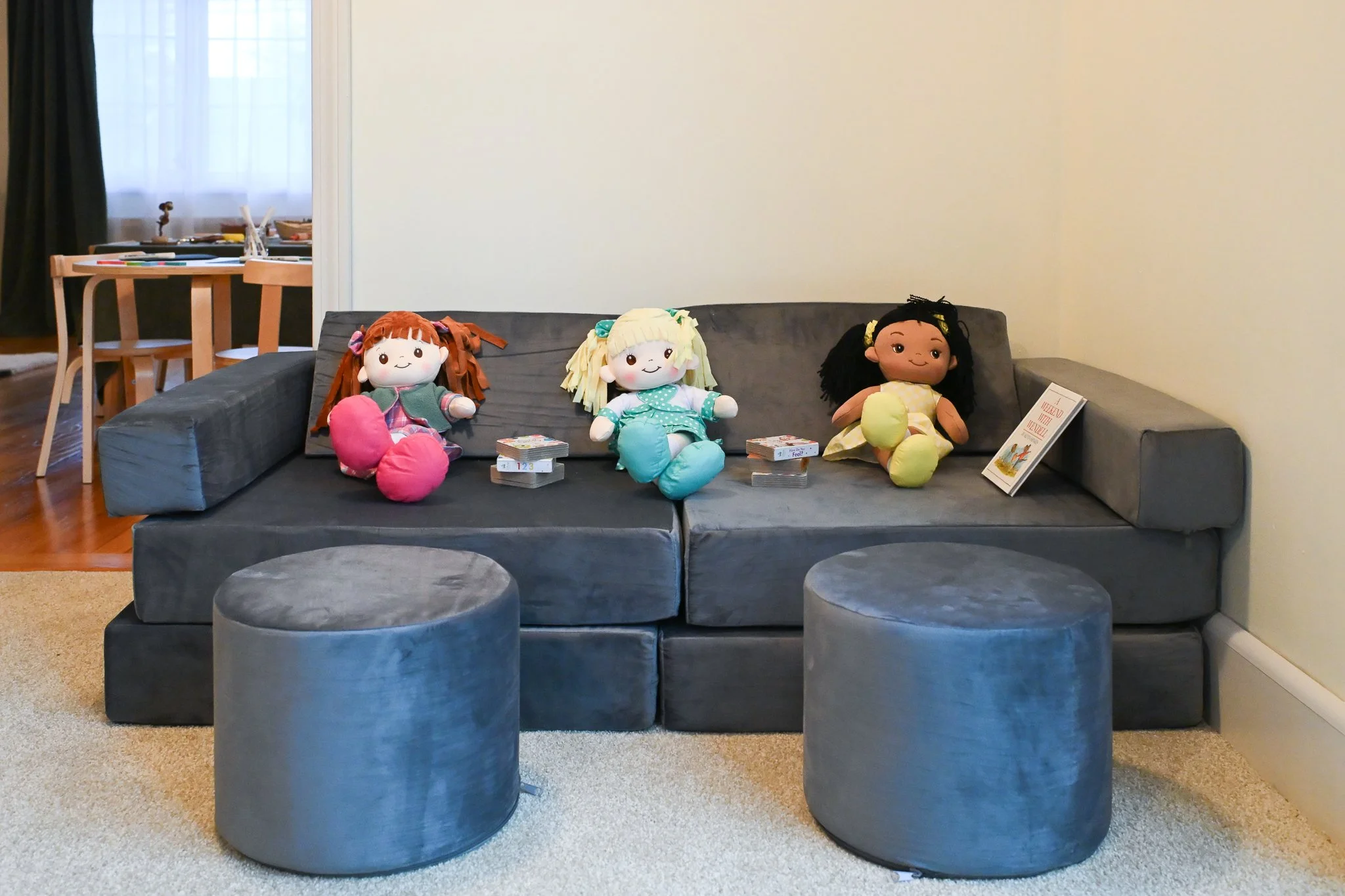 Gray sofa with three plush dolls sitting on it, and two matching ottomans in front. The background shows a dining table and chairs near a window.