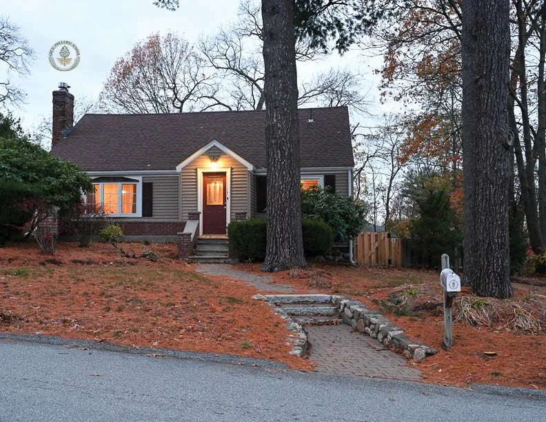 A cozy house at dusk illuminated from inside, surrounded by tall pine trees and a curved stone pathway leading to the front door.