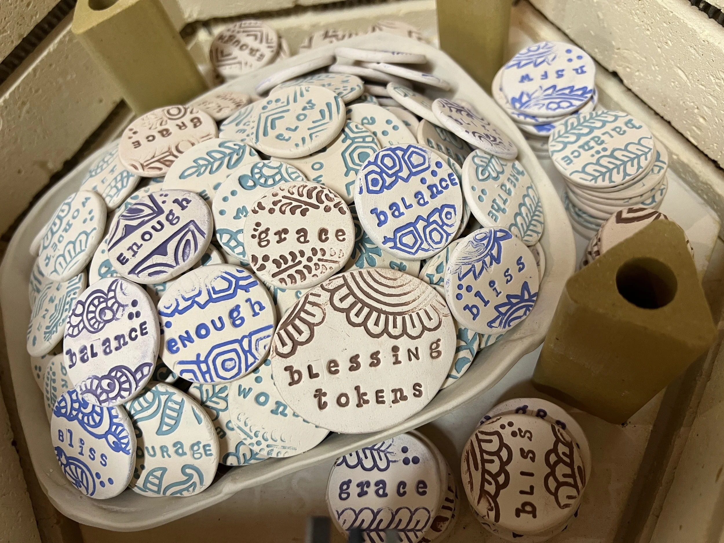 A white bowl filled with round tokens featuring words like 'bless,' 'grace,' 'balance,' 'encourage,' and 'hope,'  with hand-stamped and colored decorative designs in blue, brown, and purple ink, inside a kiln.