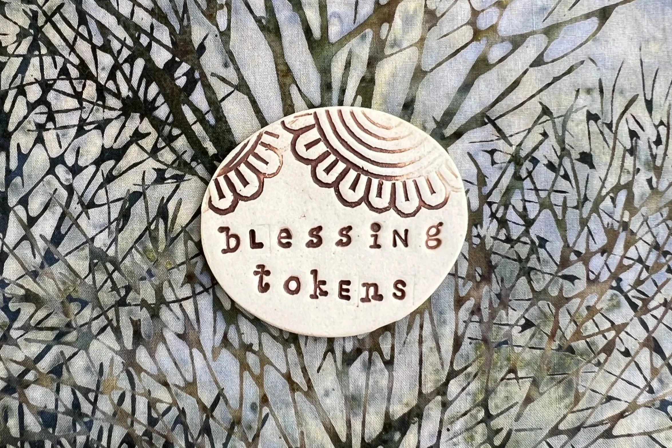 A decorative circular token with the words 'blessing tokens' engraved on it and a stylized design of leaves and rainbows above the text, placed on a fabric with a leaf pattern.