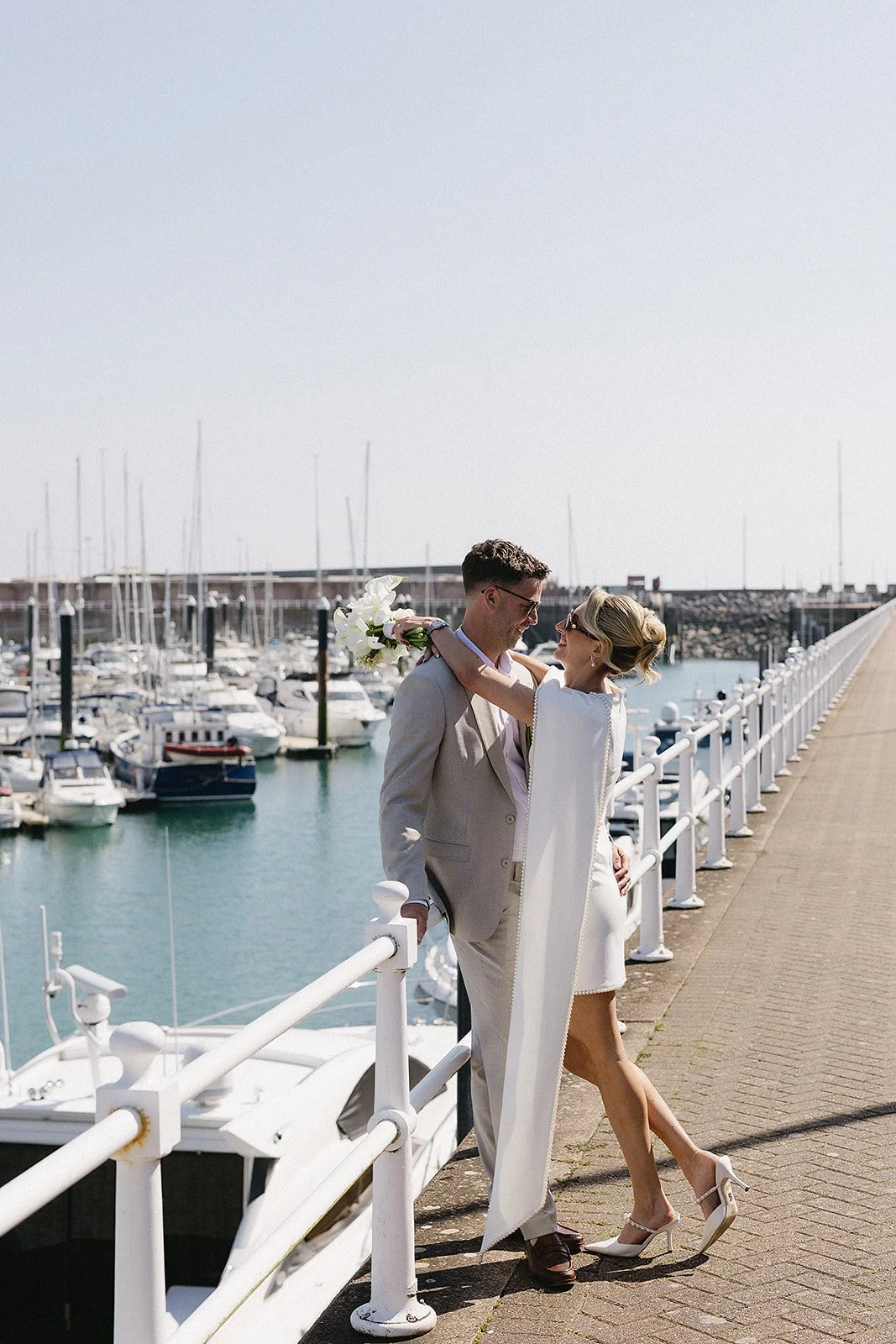 A man and woman in wedding attire celebrating by a marina with sailboats in the background. The woman is holding a bouquet and affectionately embracing the man.