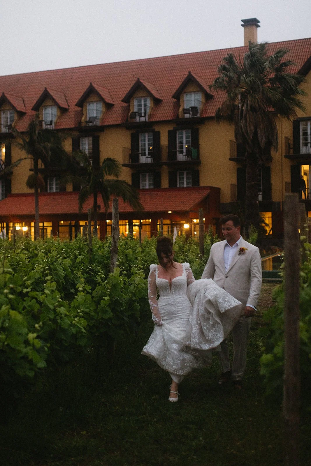 A bride and groom walking through a vineyard at sunset, with a large yellow building with red roofs and palm trees in the background.