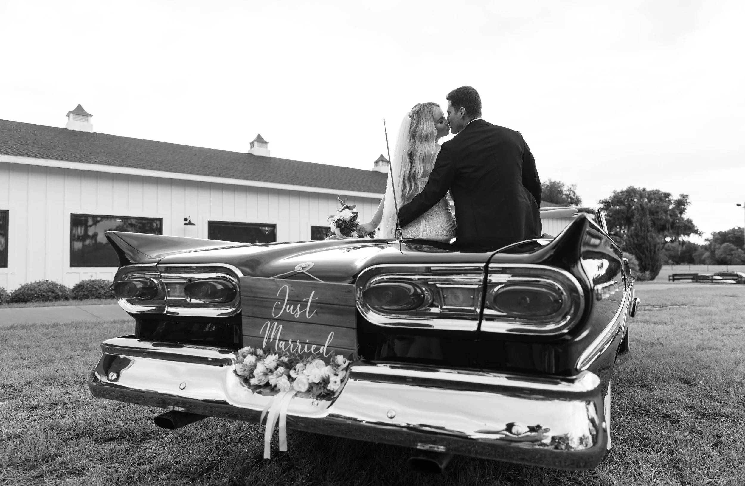 A couple dressed in wedding attire sharing a kiss on top of a vintage Cadillac with a sign reading 'Just Married' and flowers on the front bumper, outdoors near a white building.
