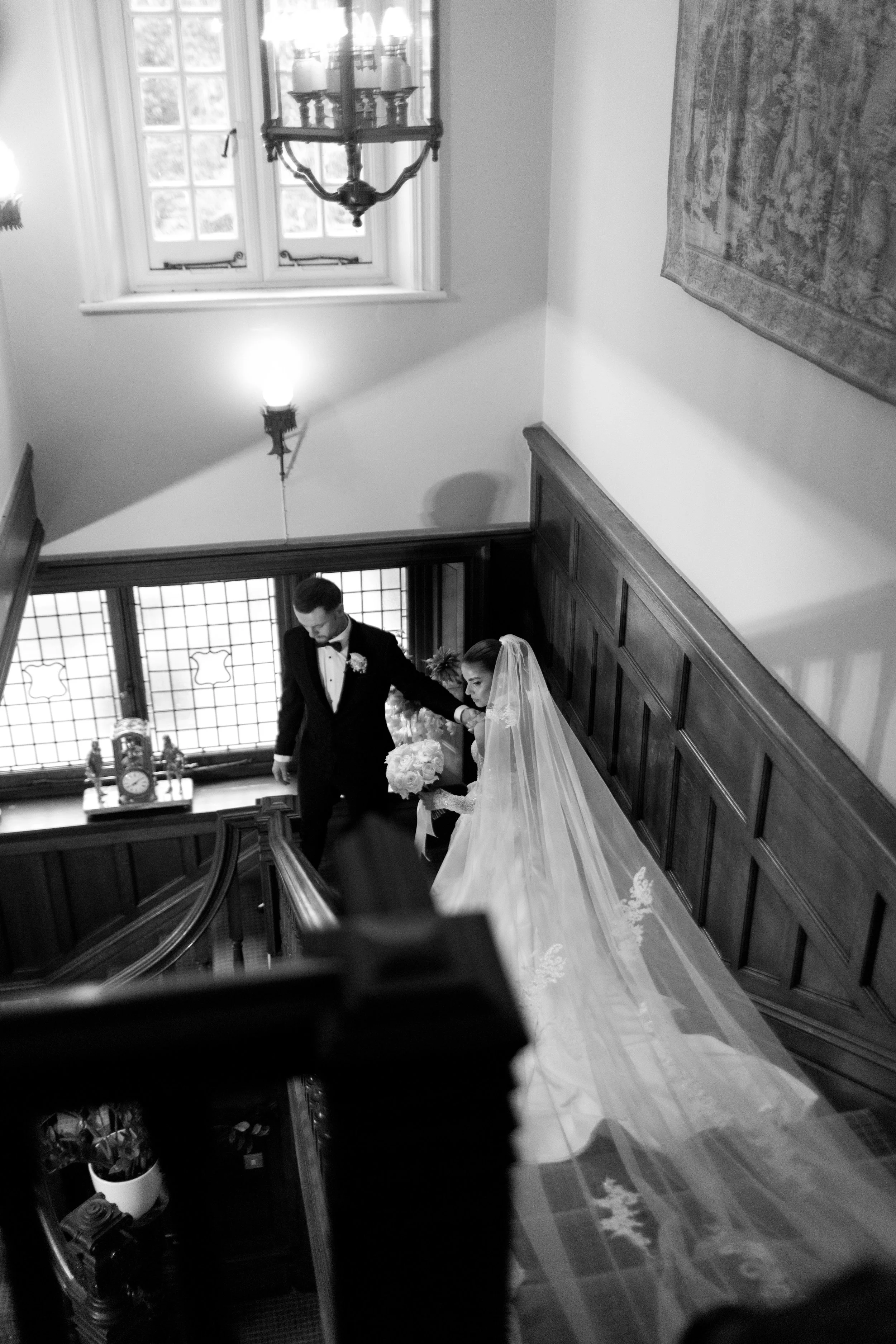 A black and white photo of a bride and groom descending a staircase during a wedding ceremony inside a historic building with wood-paneled walls and stained glass windows.