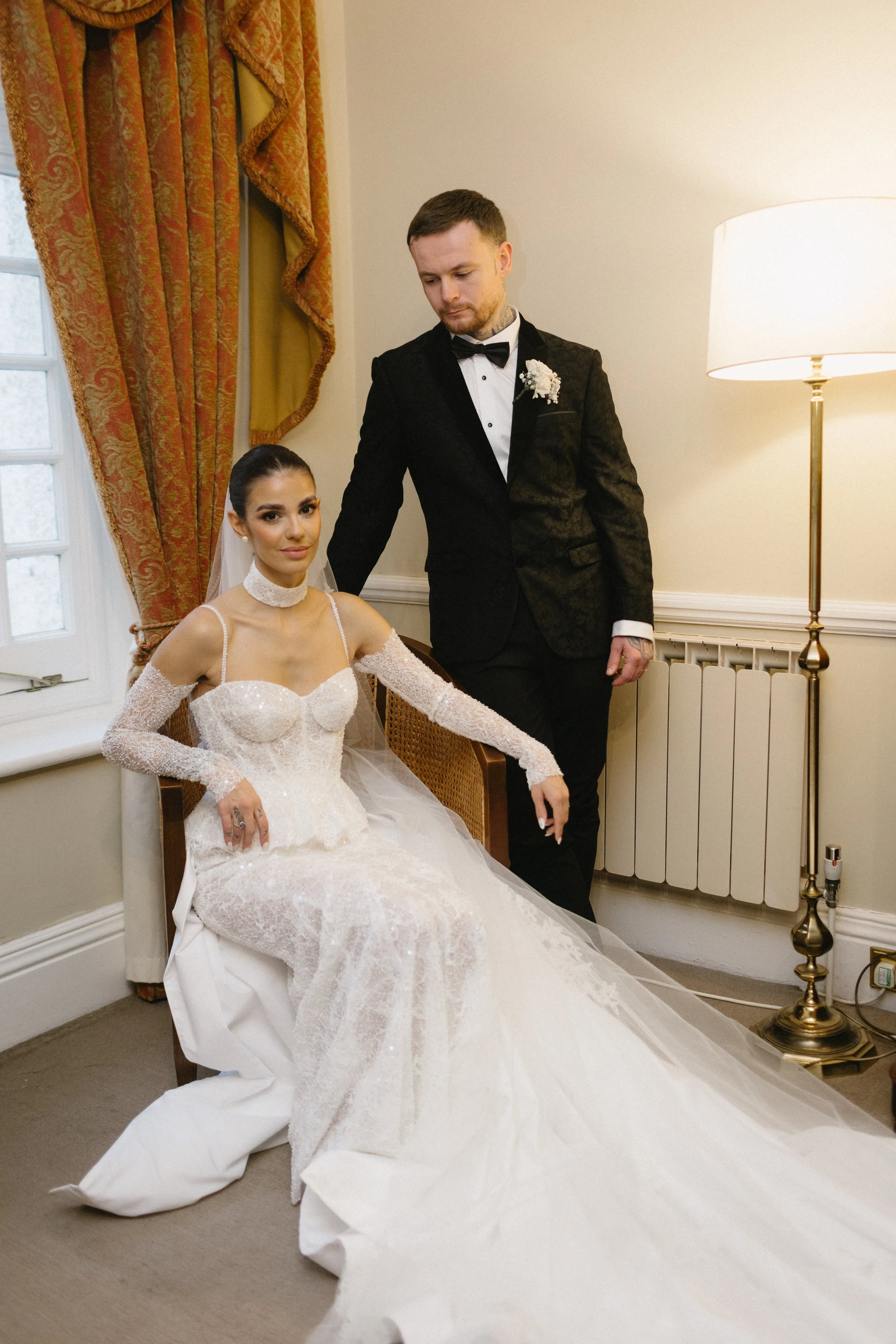 A bride in a white wedding dress sitting on a chair with her dress spread on the floor, and a groom in a black tuxedo standing beside her, touching her shoulder. They are in a room with cream-colored walls, a window with curtains, and a tall lamp.
