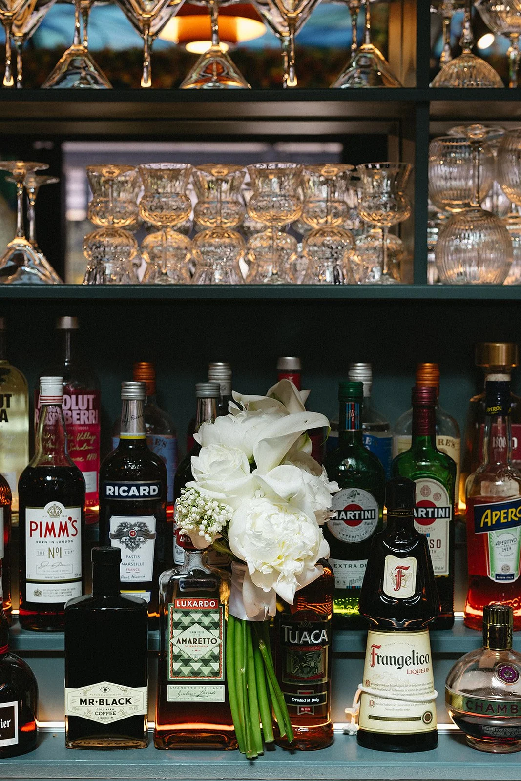 A bar shelf with various liquor bottles, a bouquet of white flowers, and upside-down wine glasses on the top shelves.