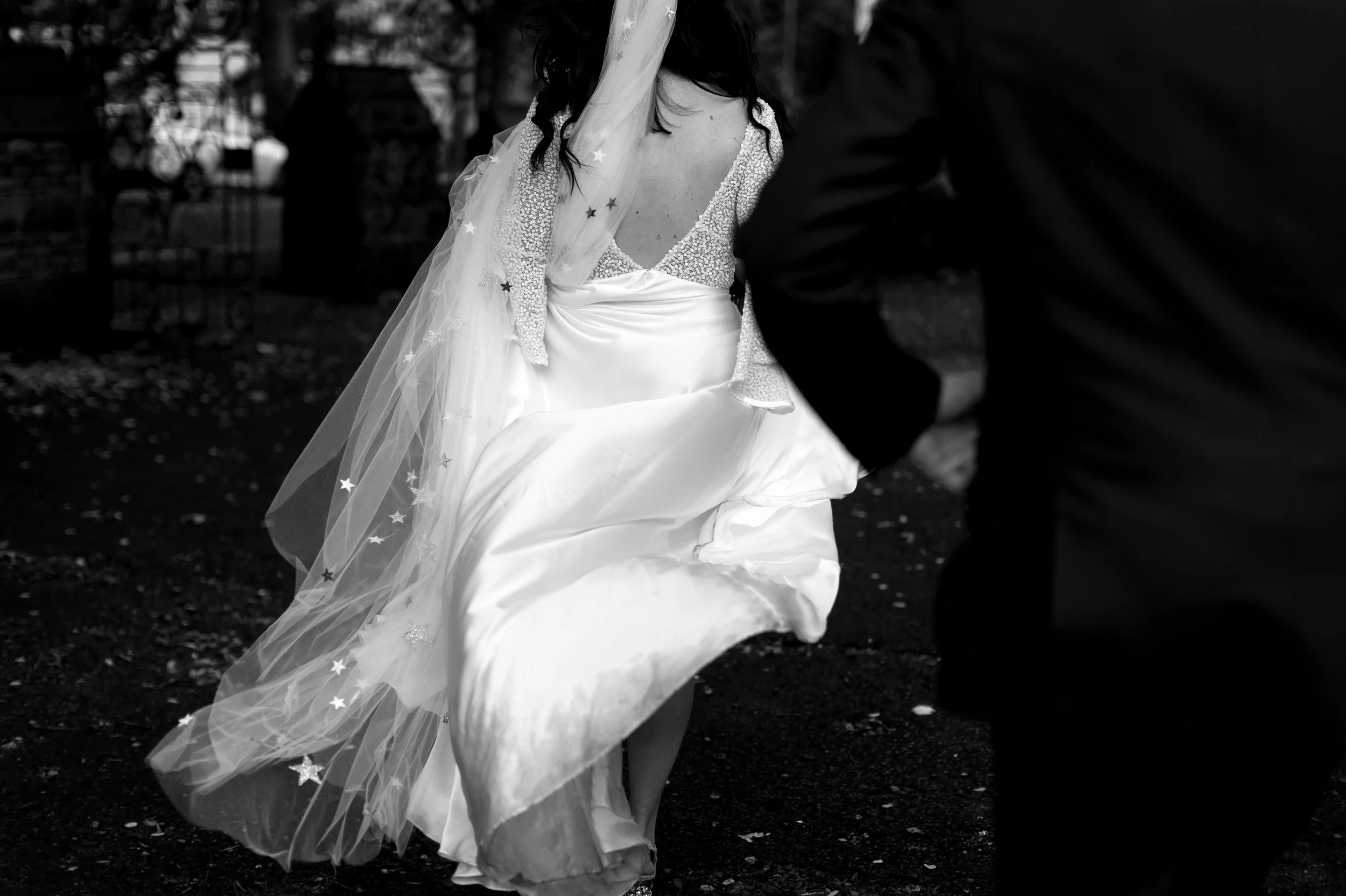 Black and white photo of a woman in a wedding dress, partially obscured, walking outdoors on grass with a veil decorated with stars.