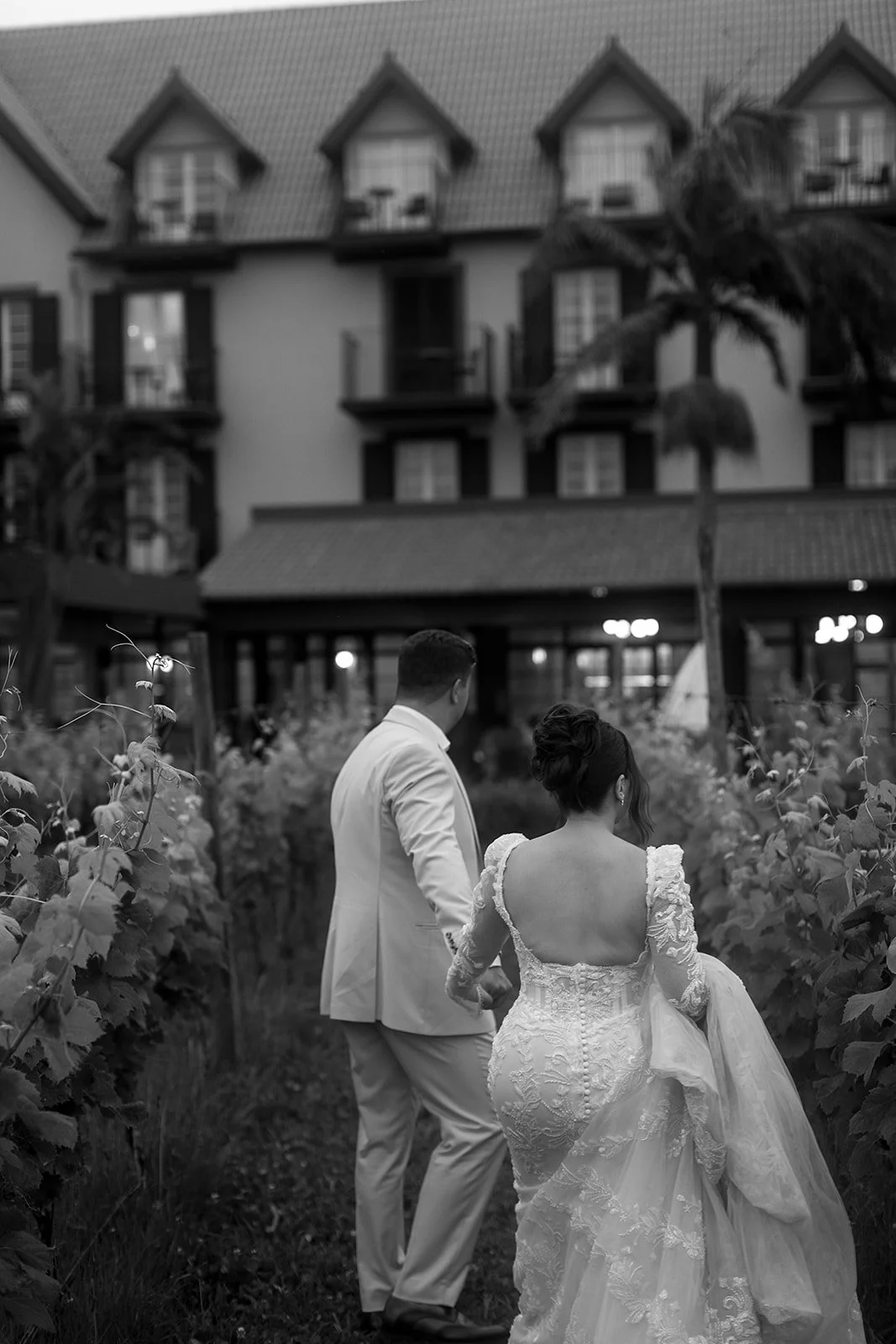 A black and white photo of a bride and groom walking through a vineyard, with a large house in the background.