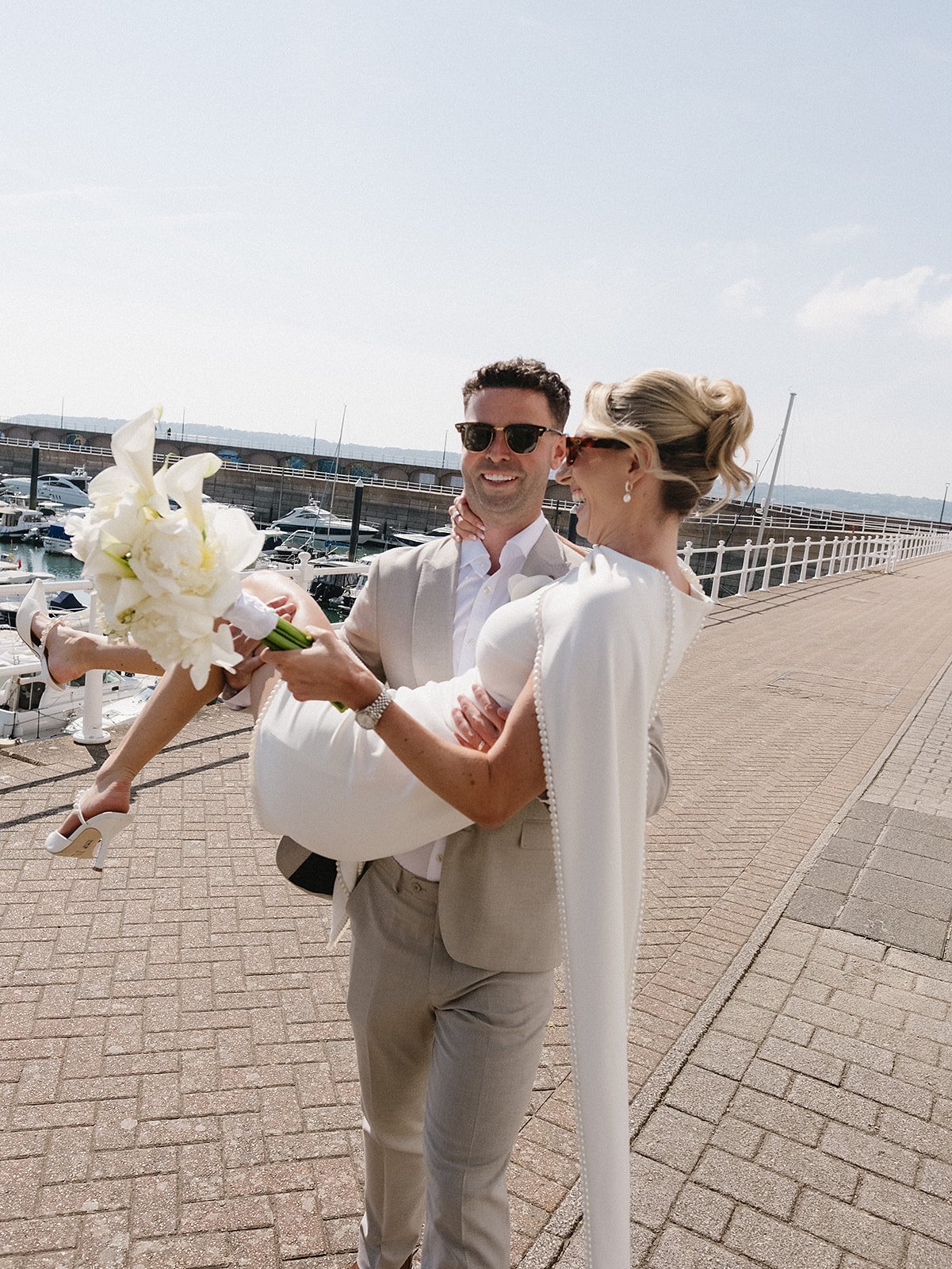 A happy couple at a marina, with the man carrying the woman and holding a bouquet of white flowers, both wearing light-colored formal attire and sunglasses, during daytime with a dock and boats in the background.