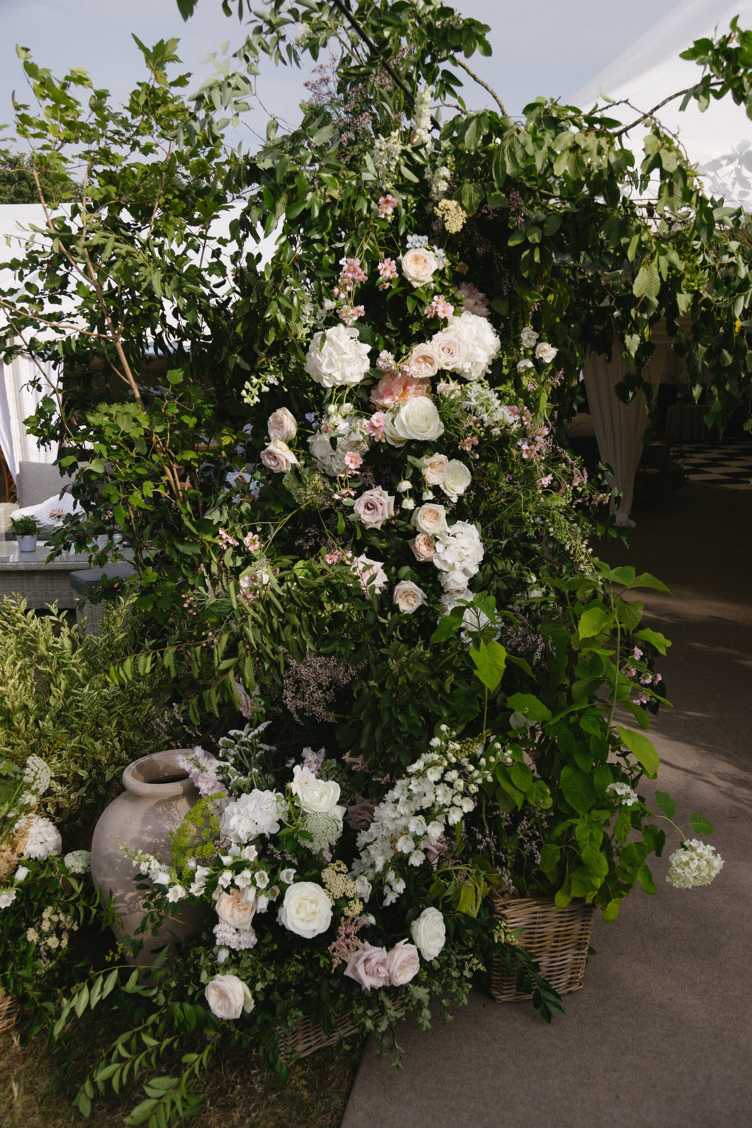 A floral arrangement featuring white and pale pink roses, hydrangeas, and other small flowers displayed in a wicker basket and a large white ceramic pot, with green foliage surrounding it, set outdoors on a paved area.
